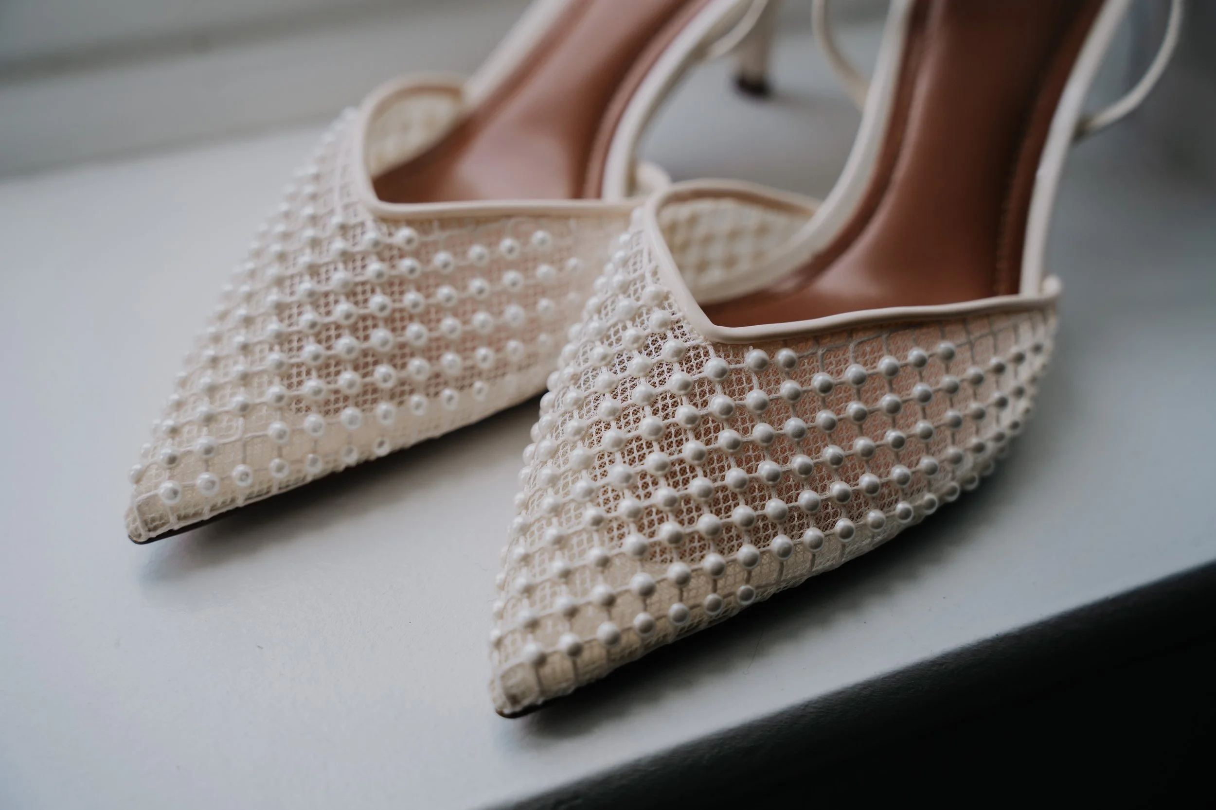 A pair of white, pointed-toe high heel shoes decorated with pearls and lace, placed on a white surface.