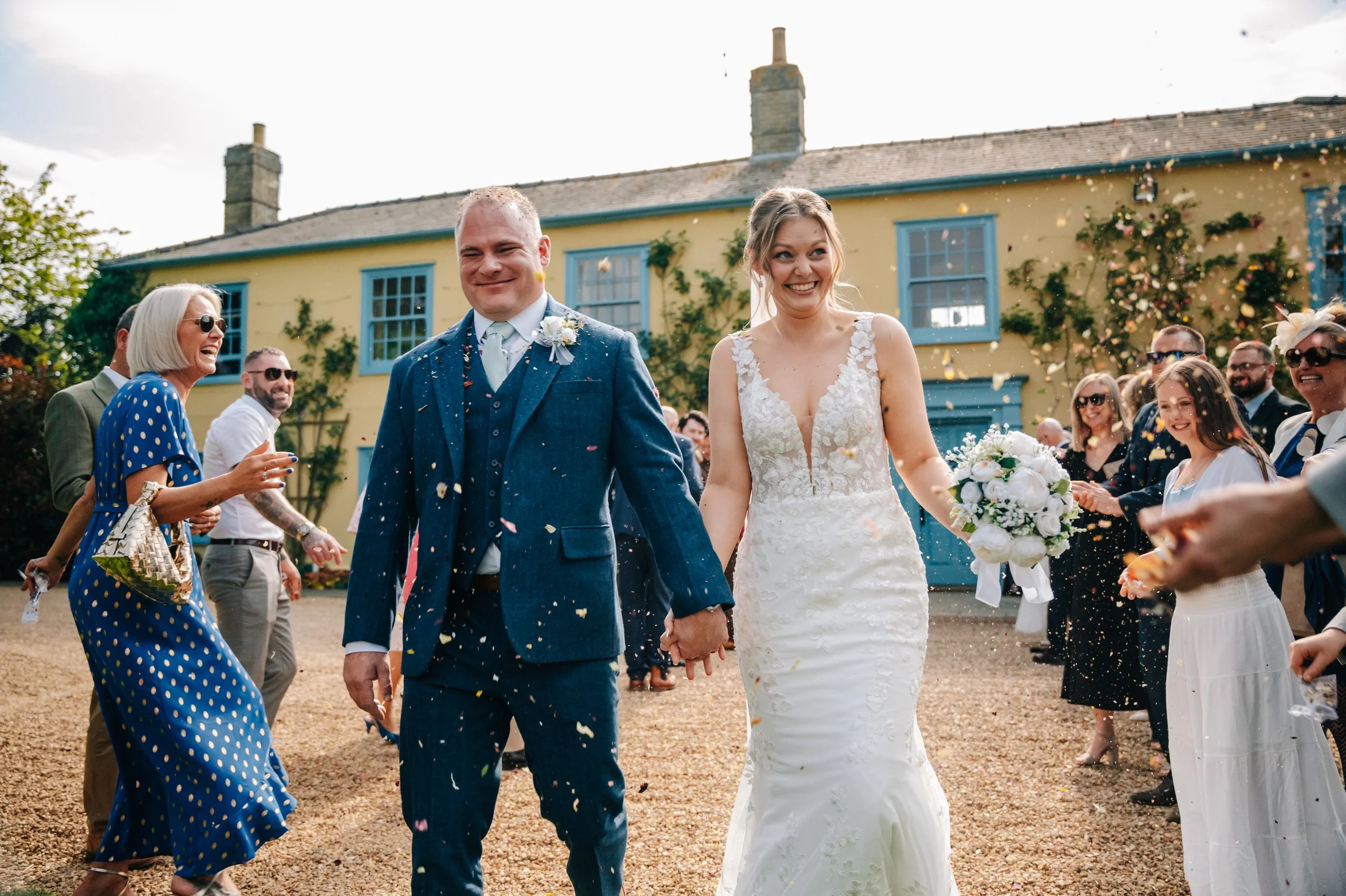 Happy newlywed couple holding hands and walking away from guests celebrating outdoors, with guests throwing confetti and smiling, in front of a yellow house with blue window frames.