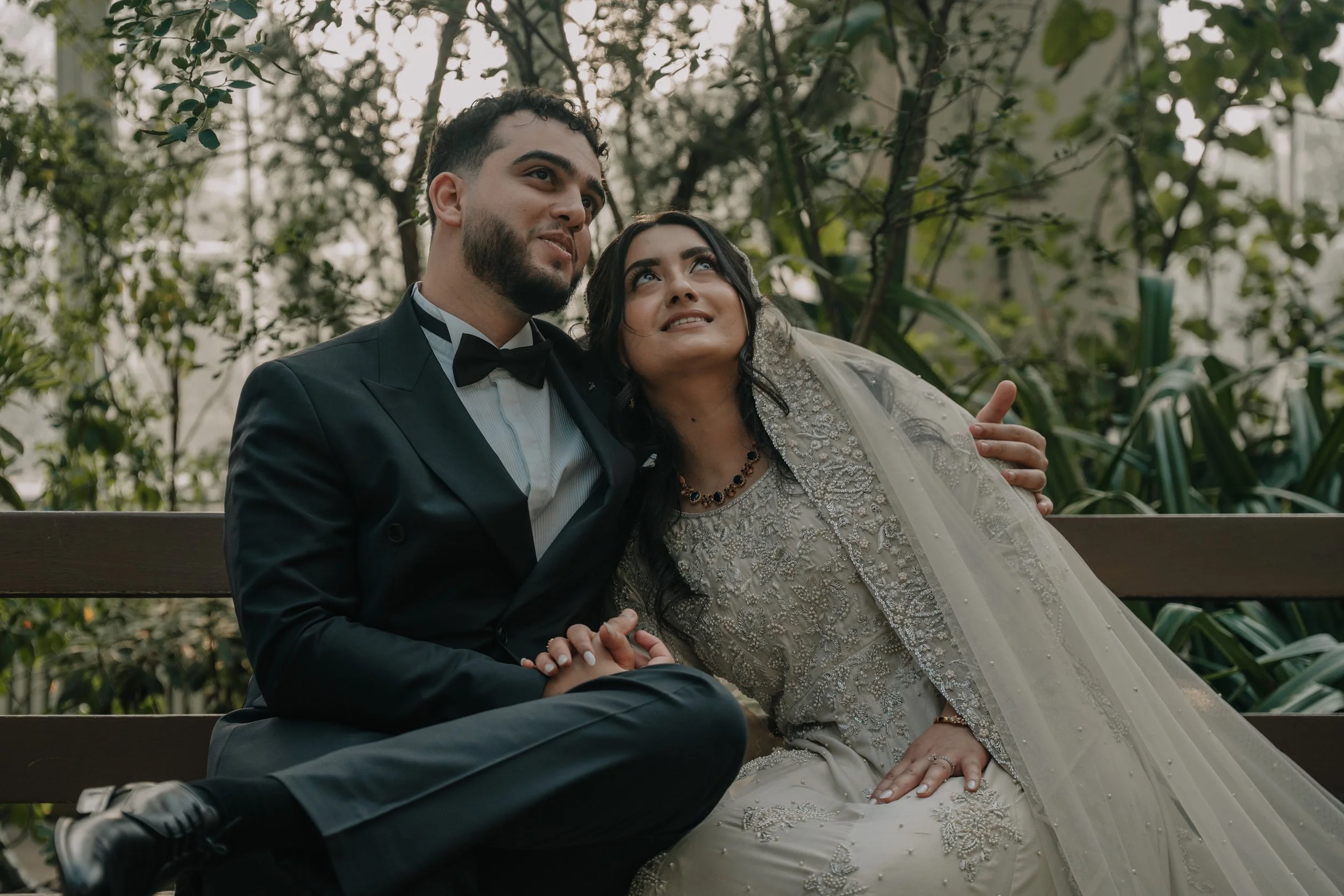 A bride and groom sitting close together on a bench outdoors, with the groom wearing a black suit and bow tie, and the bride dressed in an ornate white wedding gown with a sheer veil, surrounded by greenery.