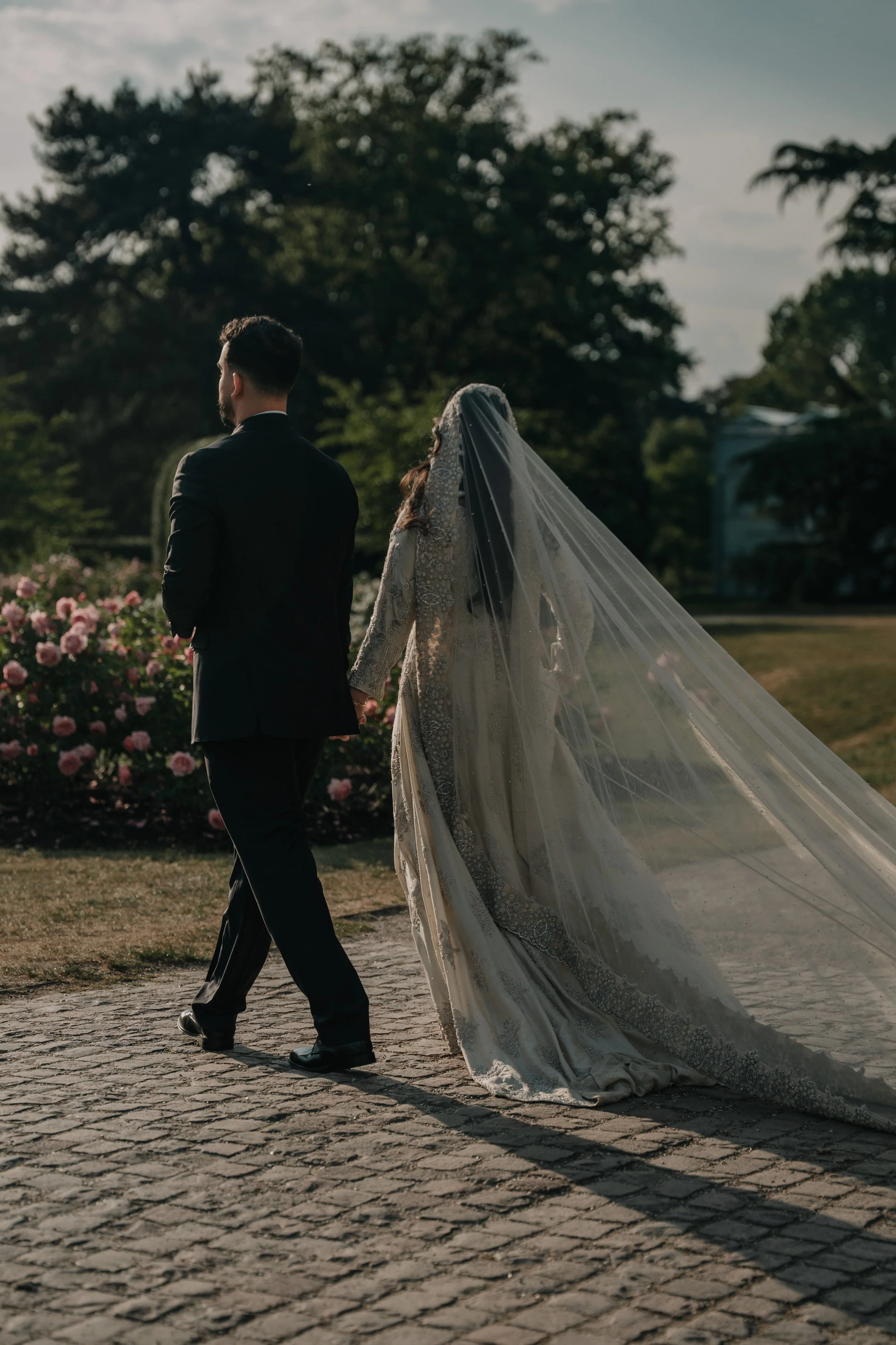 A bride and groom walking hand in hand outdoors on a cobblestone path with pink flowers and green trees in the background.