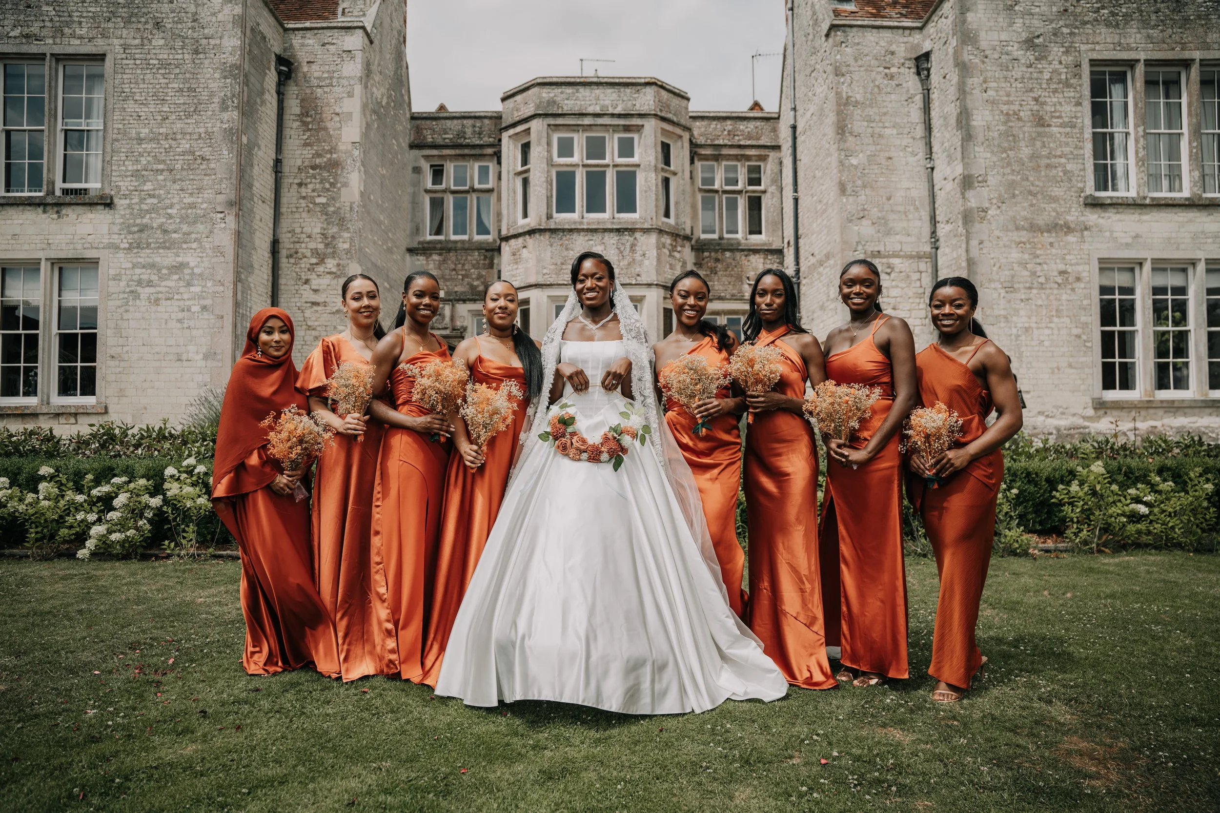 A bride in a white wedding gown with a floral belt and veil, standing among nine bridesmaids dressed in matching rust-orange dresses, outside in front of a historic stone building with multiple windows, during daytime.