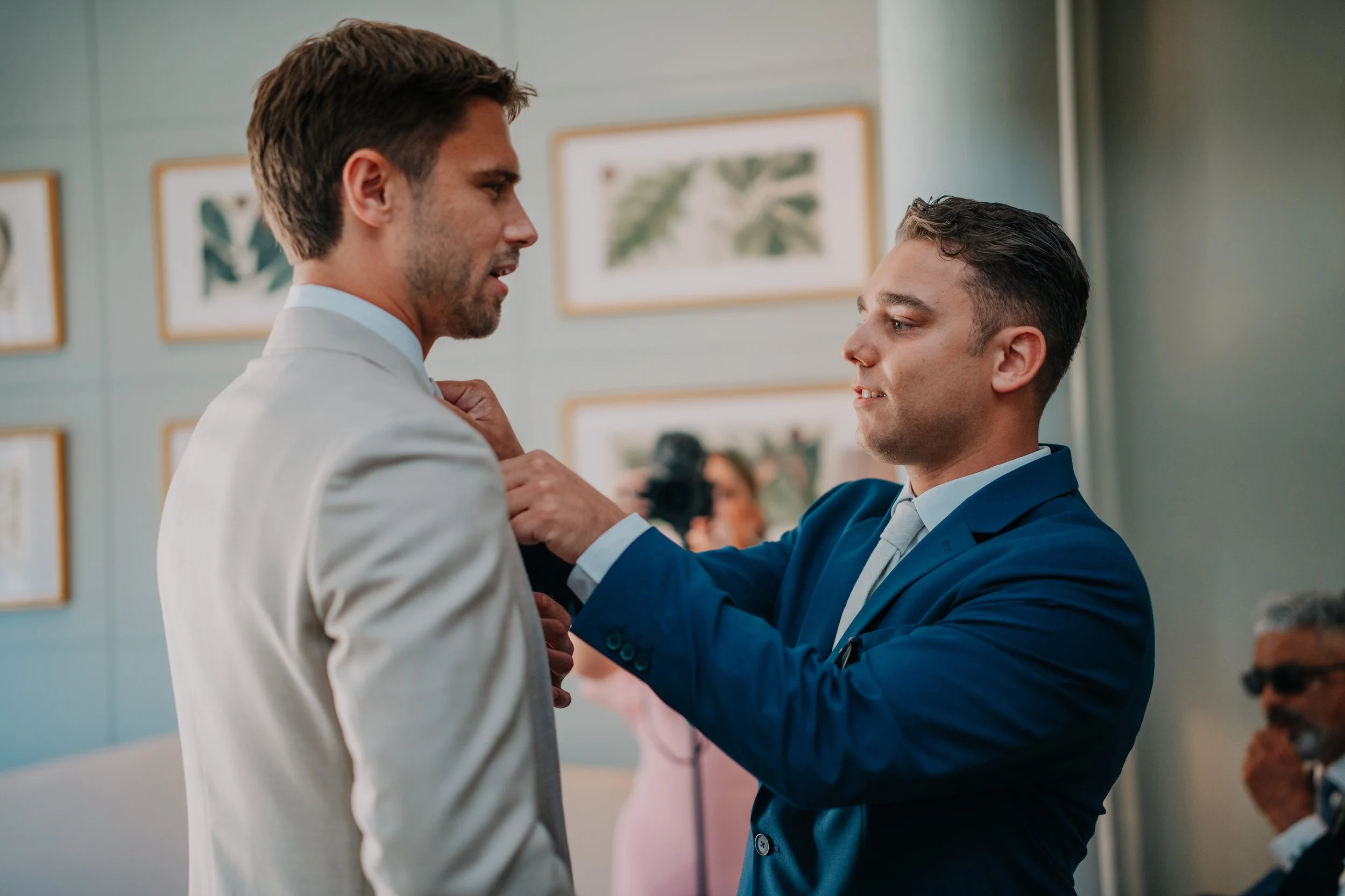 Two men, one in a white suit and the other in a blue suit, are adjusting the other's suit jacket in a room with artwork on the wall.