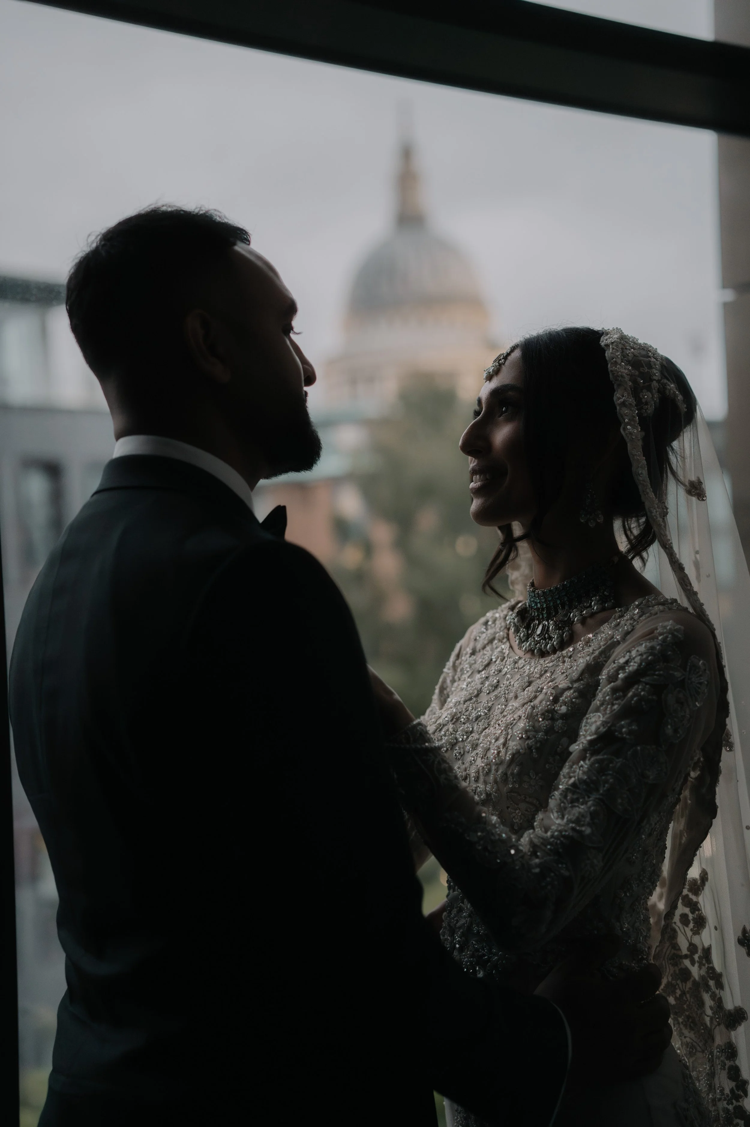 Silhouette of a bride and groom holding hands indoors, with blurred dome building in the background through a window.