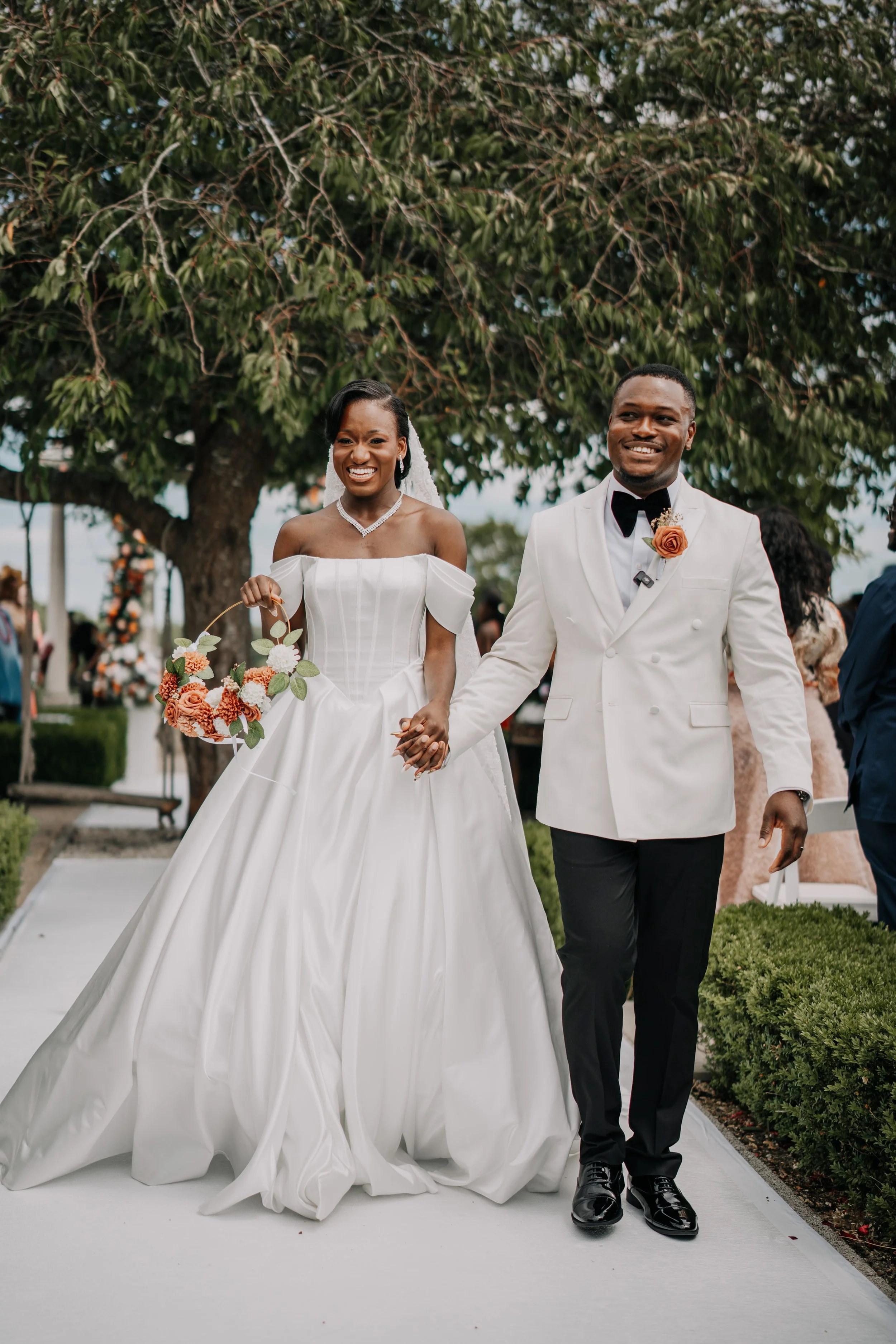 A newlywed couple walking hand in hand at an outdoor wedding ceremony. The bride is wearing a white off-shoulder gown and holding a bouquet, while the groom is dressed in a white tuxedo jacket, black pants, and a black bow tie.