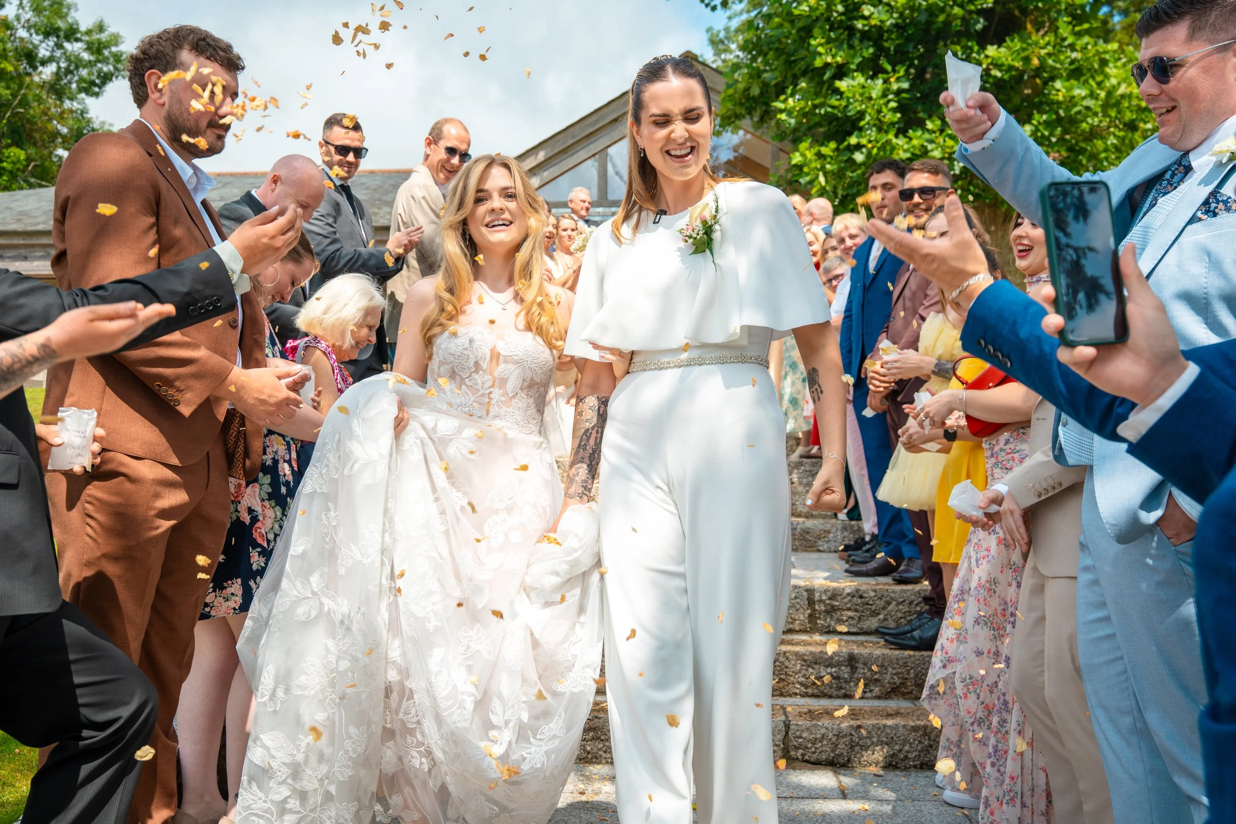 Women in wedding attire walking down steps while surrounded by friends and family, celebrating with confetti and smiles at an outdoor wedding.