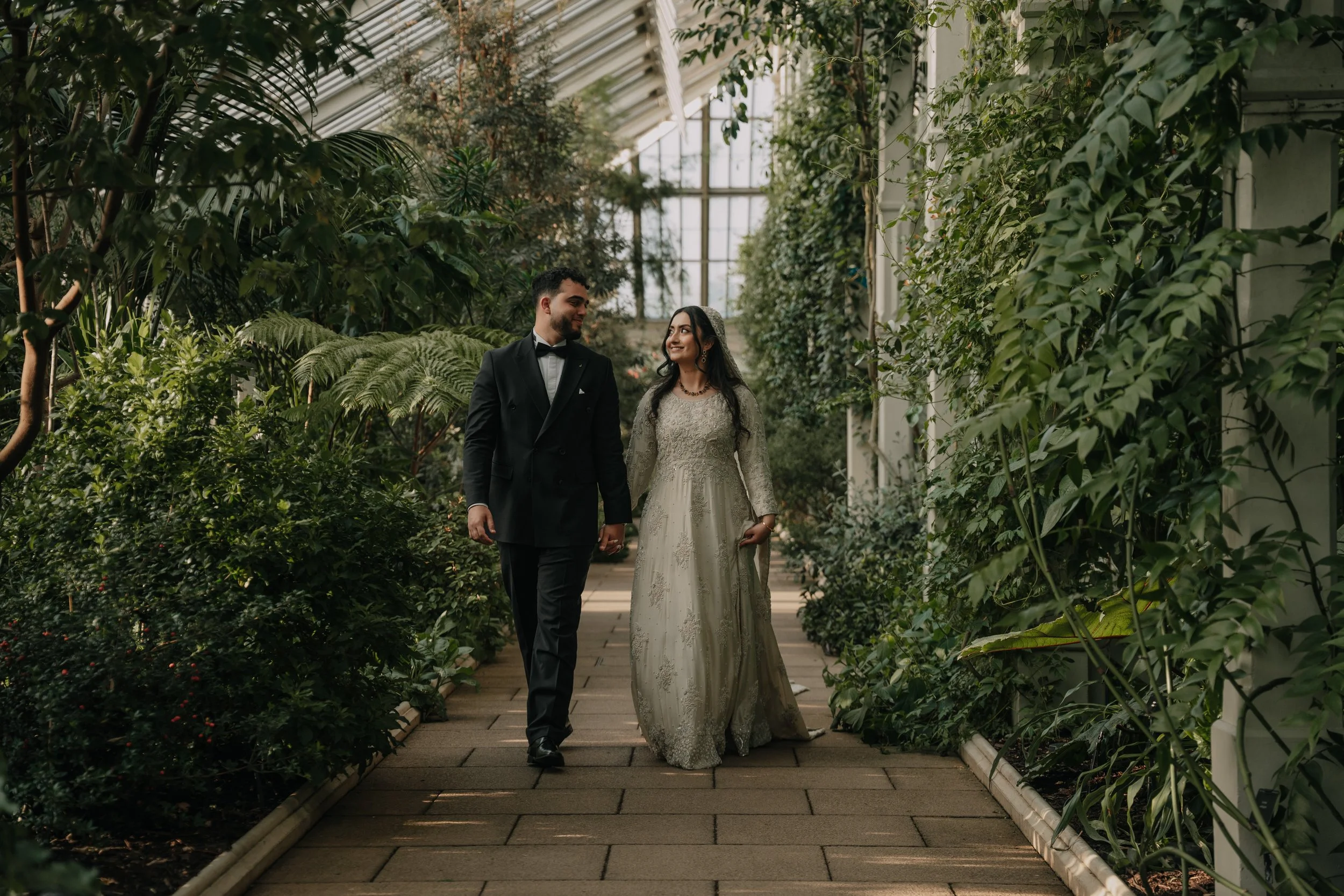 A couple in wedding attire walking hand in hand through a lush greenhouse with tall green plants and a glass ceiling.