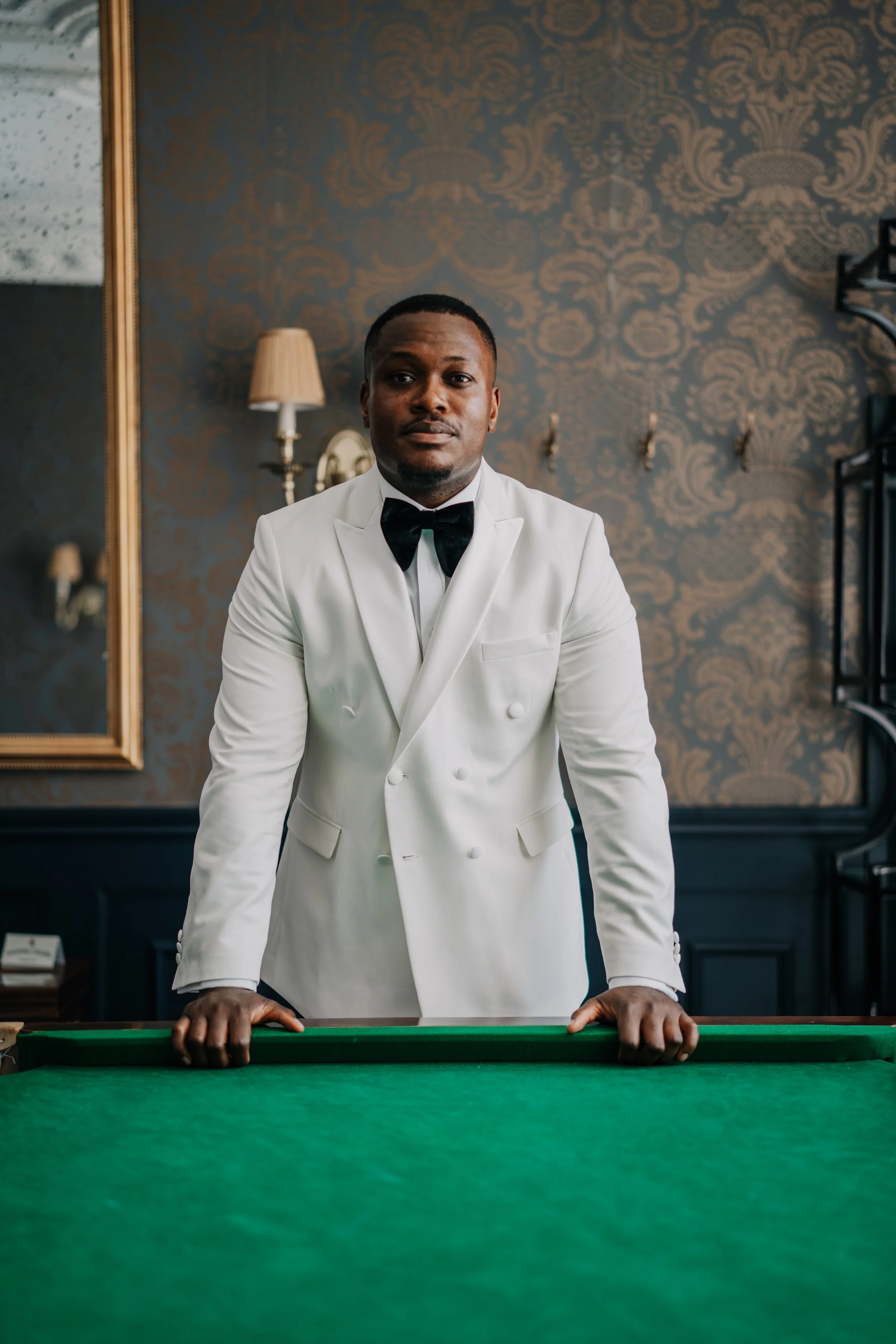 A man in a white tuxedo with a black bow tie standing behind a green felt billiard table, indoors with decorative wallpaper and a mirror to his left.