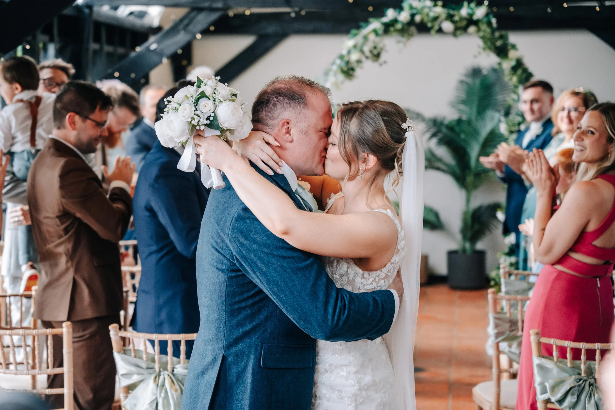 A bride and groom kiss during their wedding ceremony, surrounded by friends and family in a decorated indoor venue.