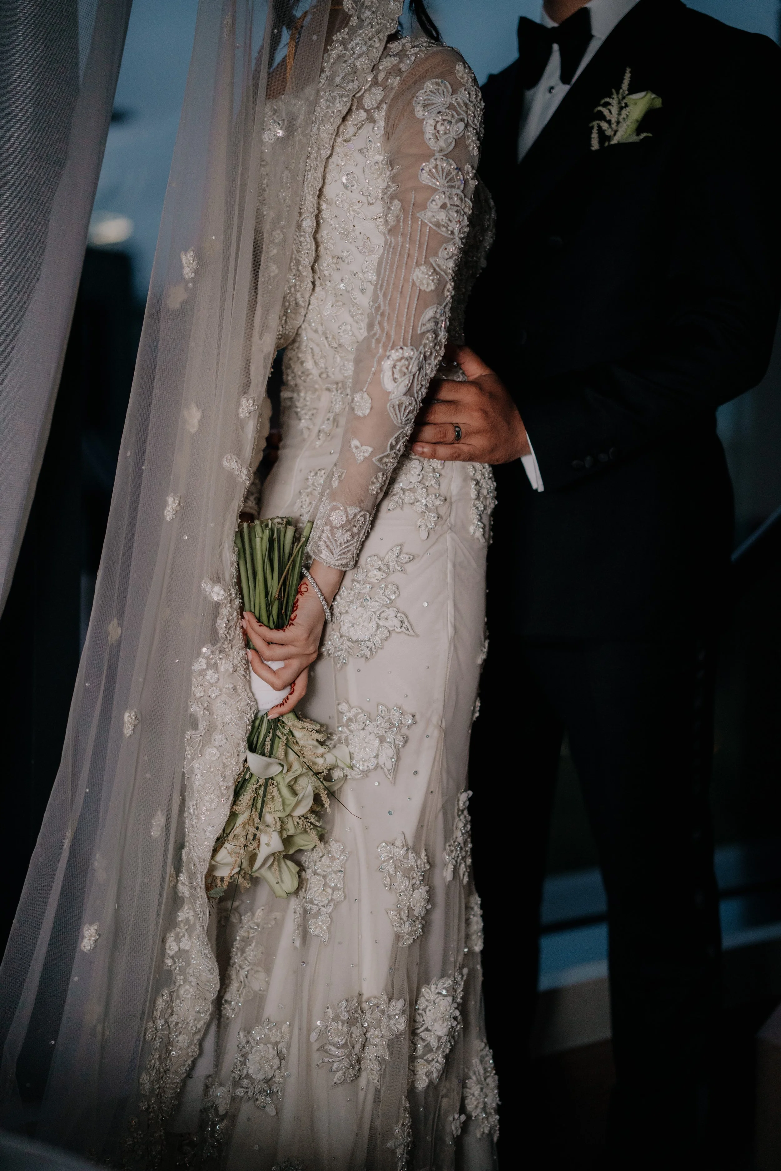 Close-up of a bride in an intricately embroidered wedding gown holding a bouquet, standing next to a groom in a black tuxedo with boutonniere.