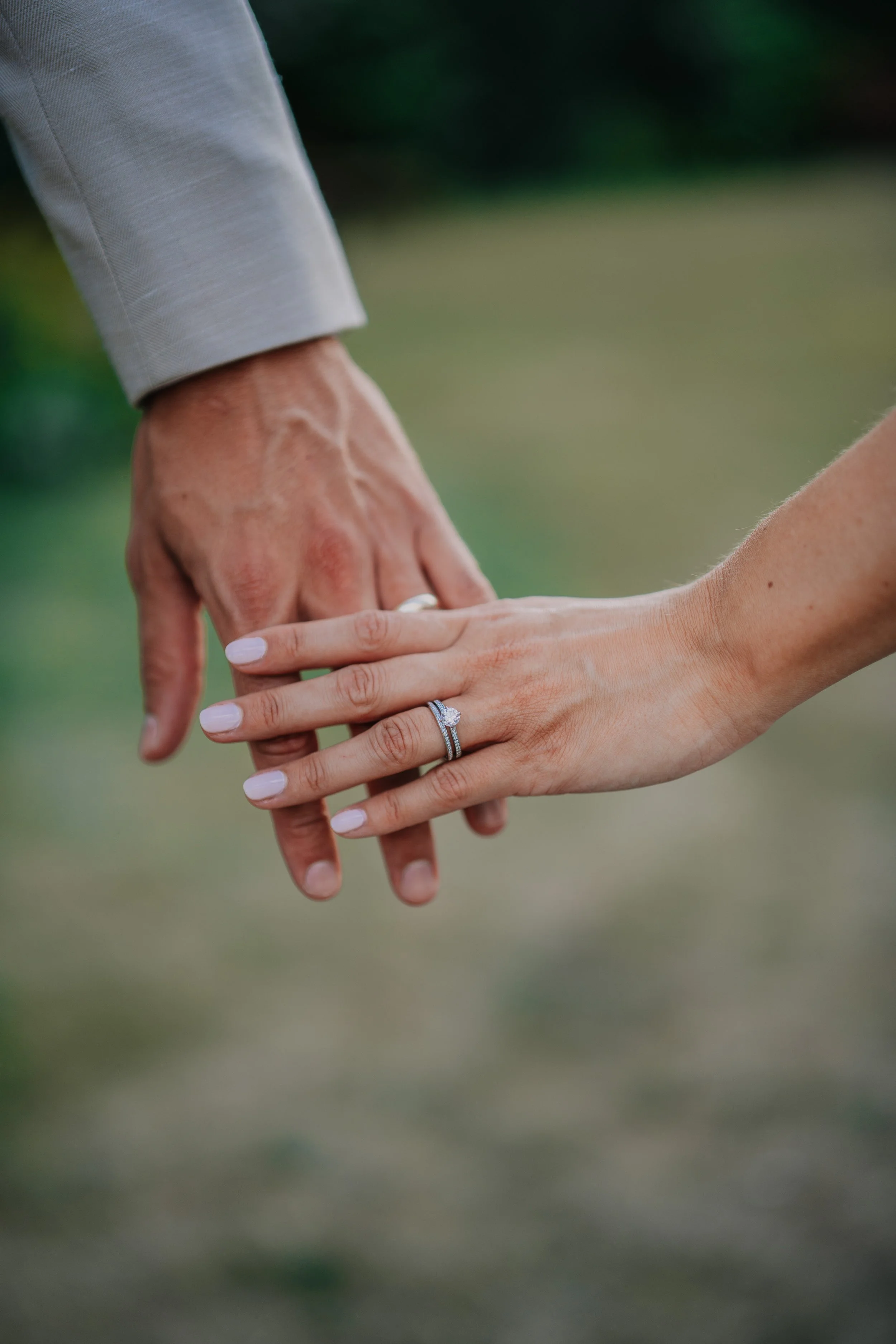 Close-up of a man and woman holding hands, the woman's hand displays a wedding ring. The background is blurred green outdoor scenery.
