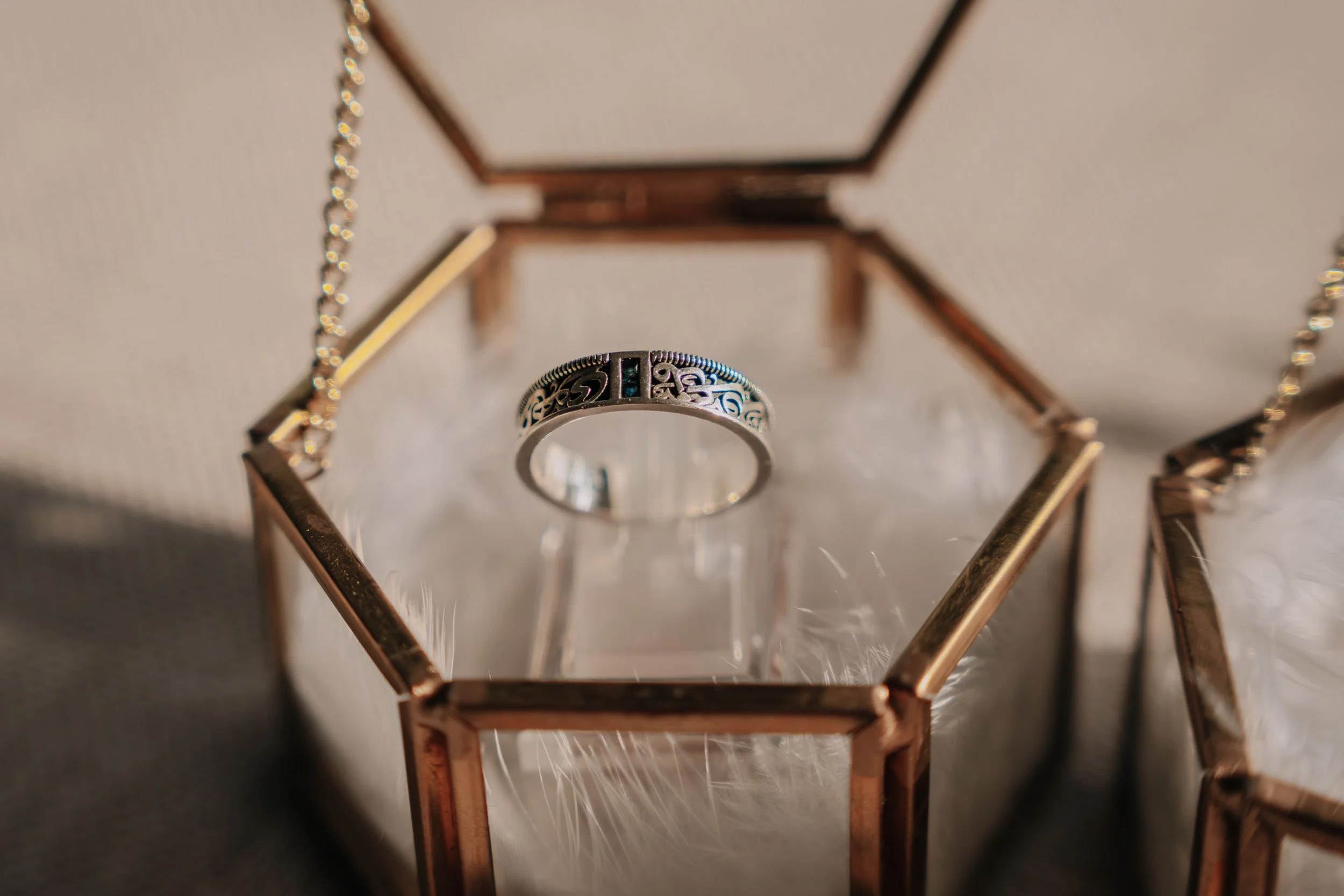 A silver ring with intricate design and small blue gemstone, displayed inside a glass and metal jewelry box.