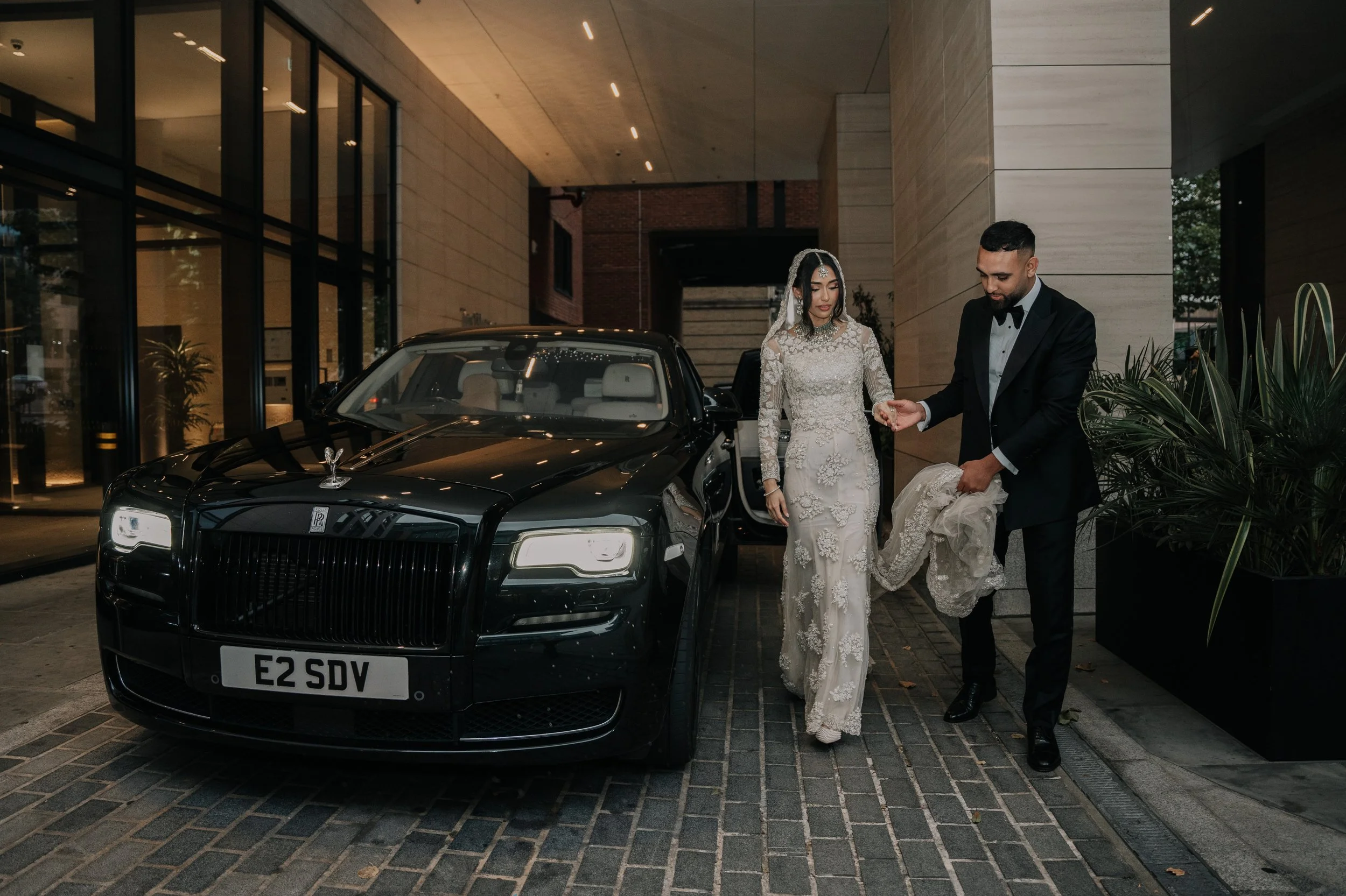 A newlywed couple, a bride in a white lace gown and veil and a groom in a black tuxedo, are outside a building near a black Rolls Royce car. The groom is helping the bride with her dress as they prepare to leave.