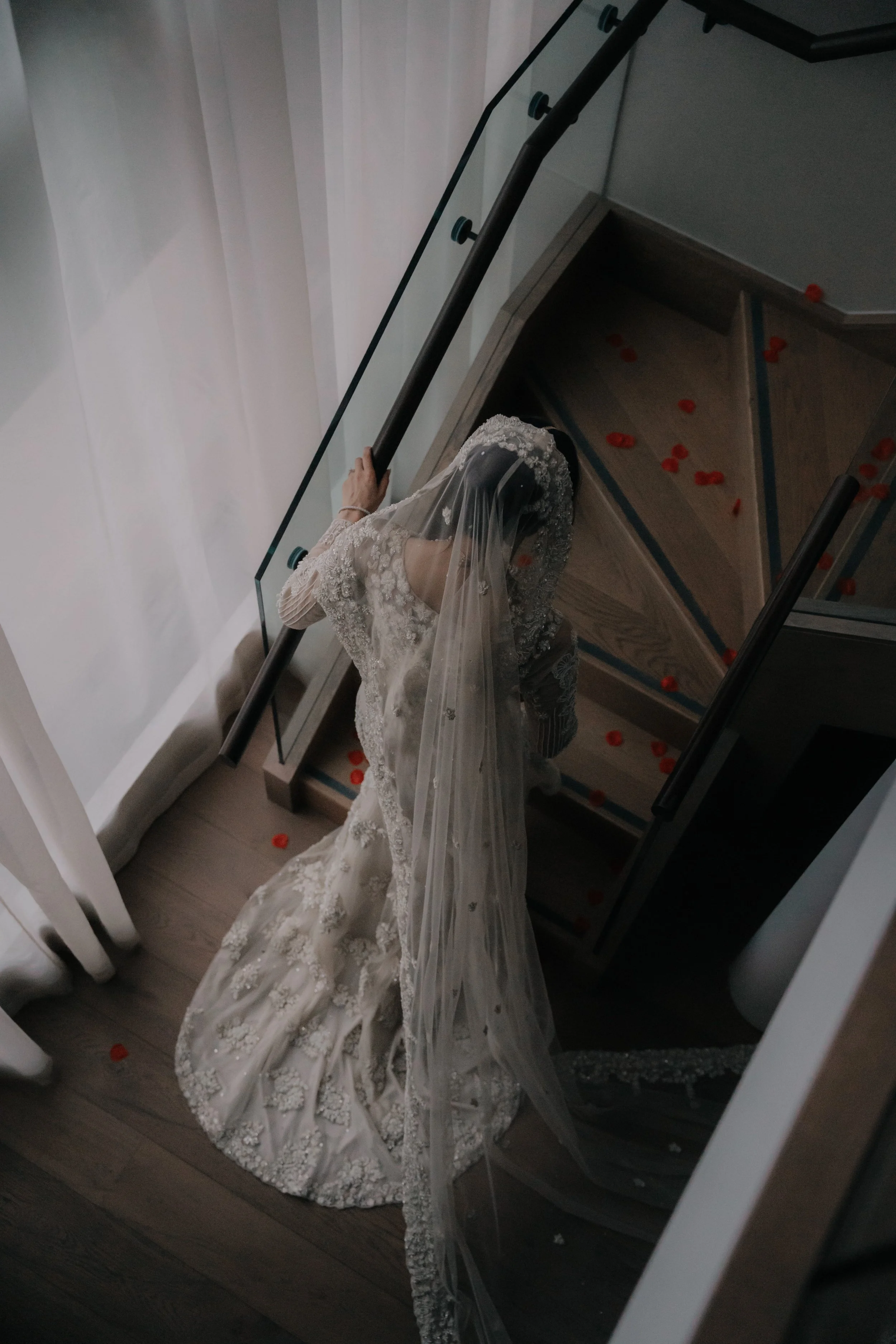 A bride in a long, detailed wedding gown with lace and embroidery, wearing a veil, descending a staircase with red flower petals scattered on the steps.