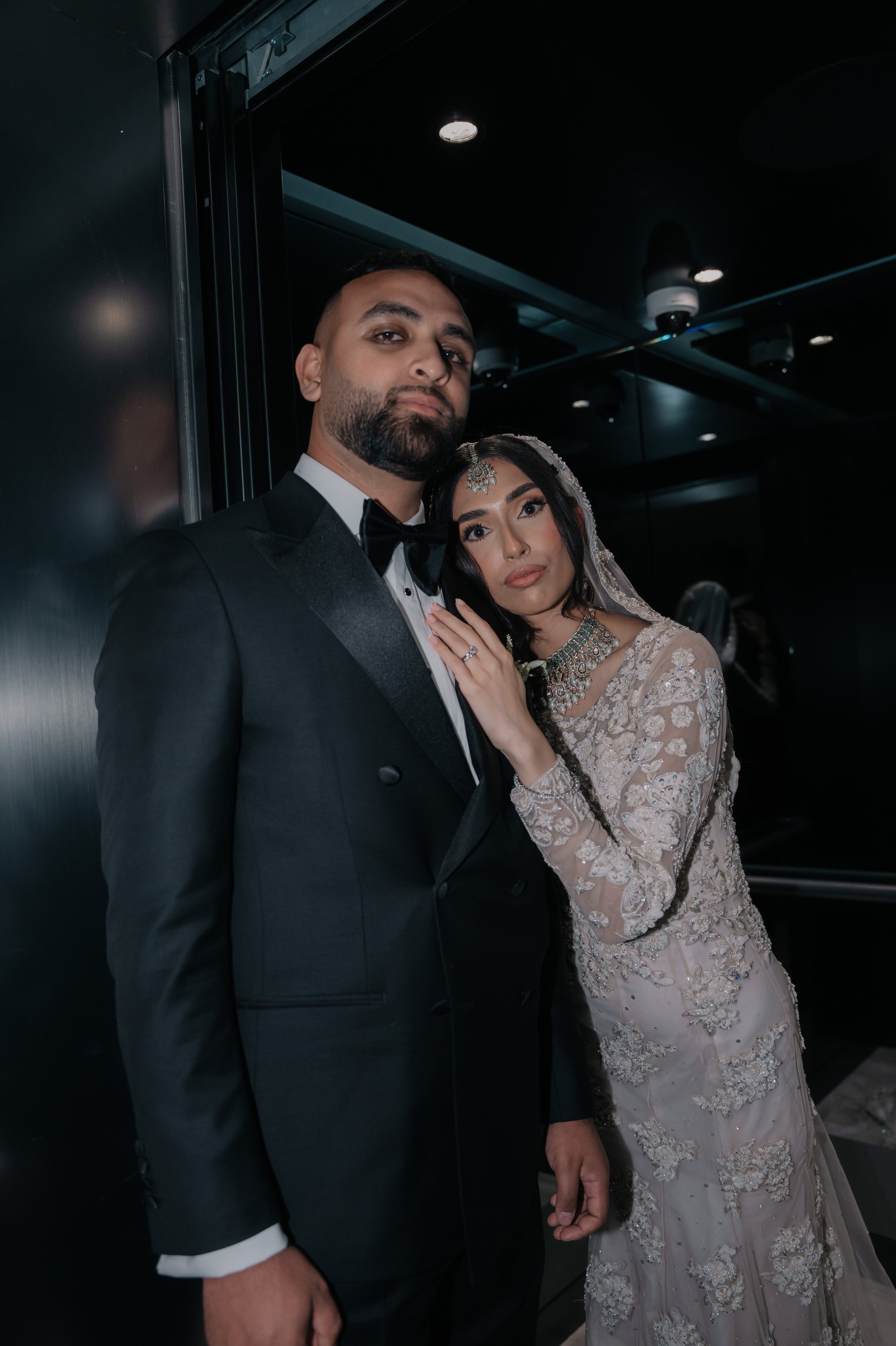 A bride and groom dressed in formal wedding attire standing close together in an elevator with dark, reflective walls and ceiling.