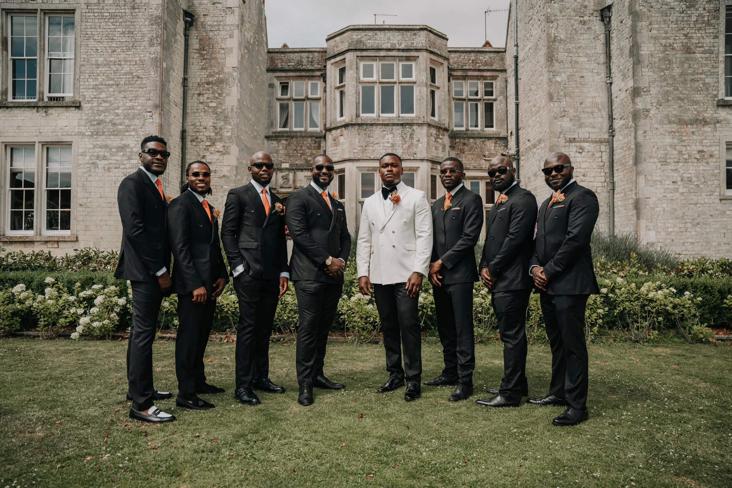 A group of nine men dressed in formal suits, with one man in the center wearing a white tuxedo, standing outdoors in front of a historic stone building with garden. The men are wearing dark suits with orange accents, and the man in white has a bouton
