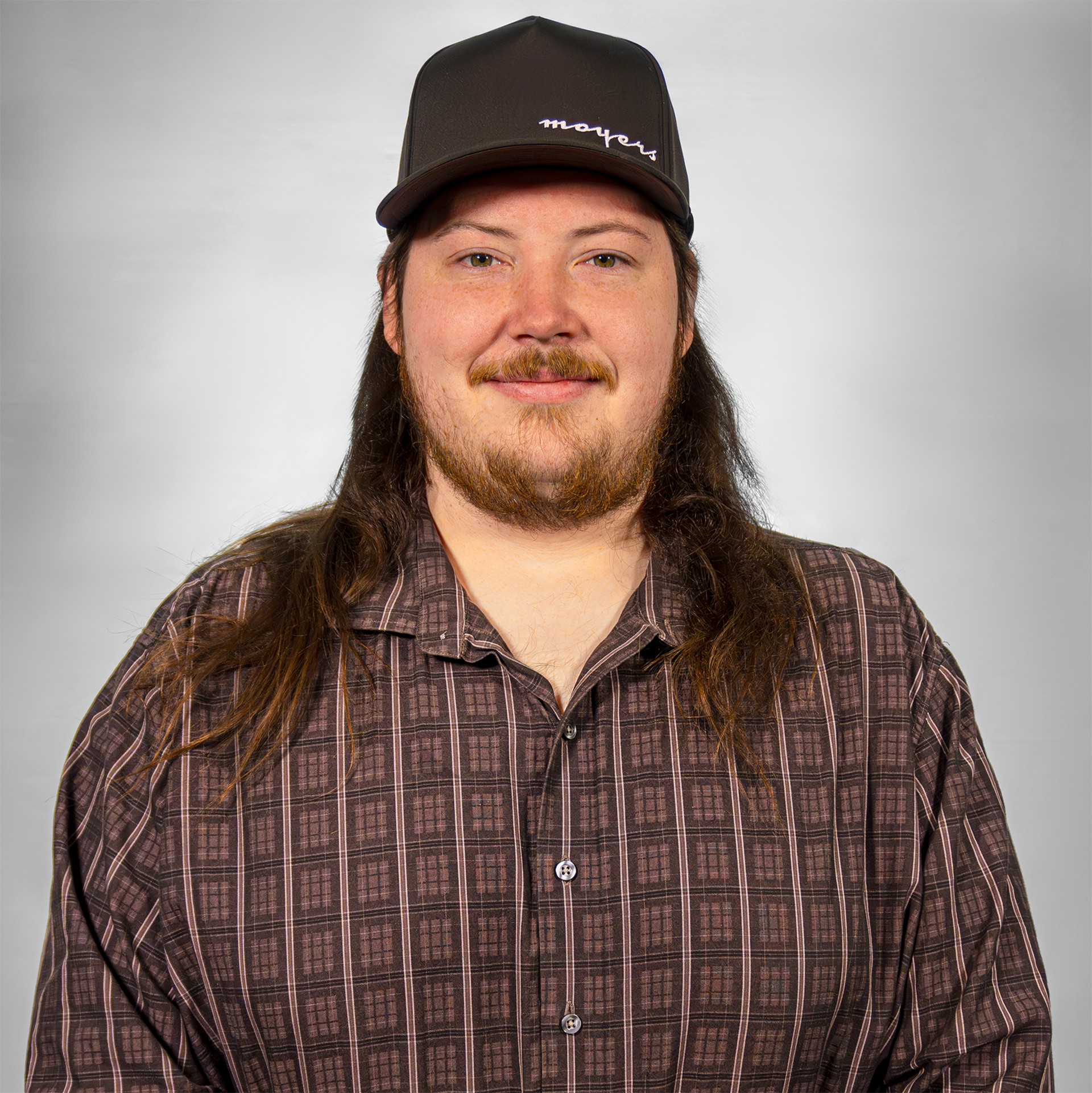 Man wearing a checkered shirt and a black cap, smiling against a gray background.