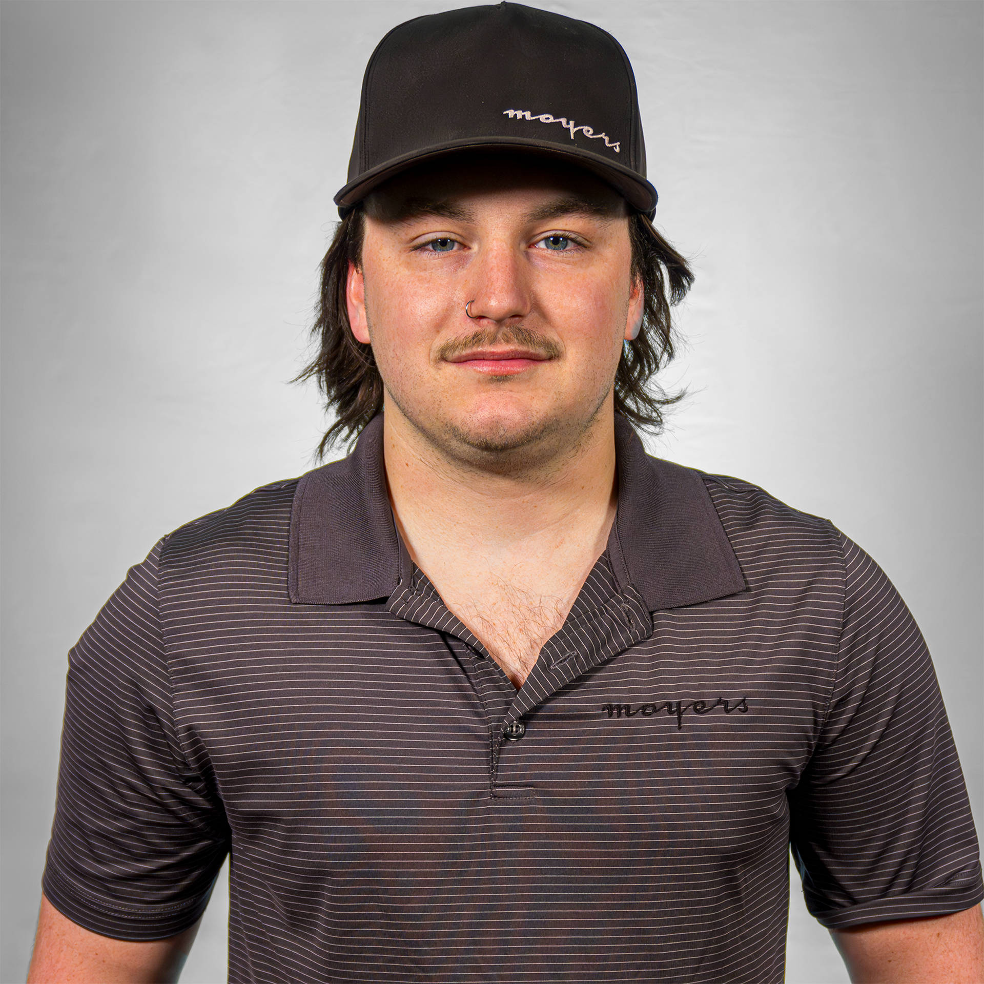 A man with a mustache wearing a black cap and a striped polo shirt in front of a plain background.