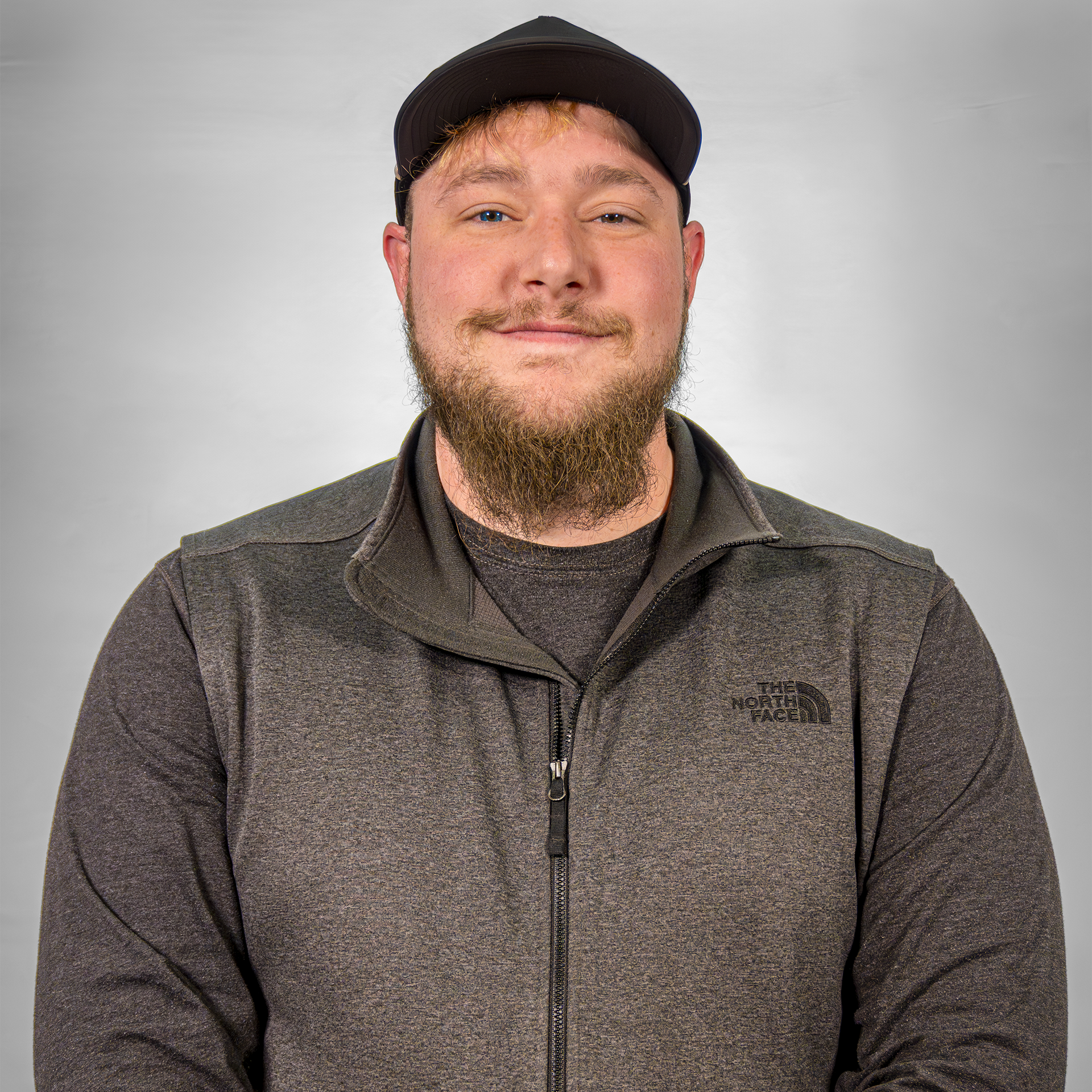 Man in a black cap and gray zip-up jacket with a beard, standing against a plain background.
