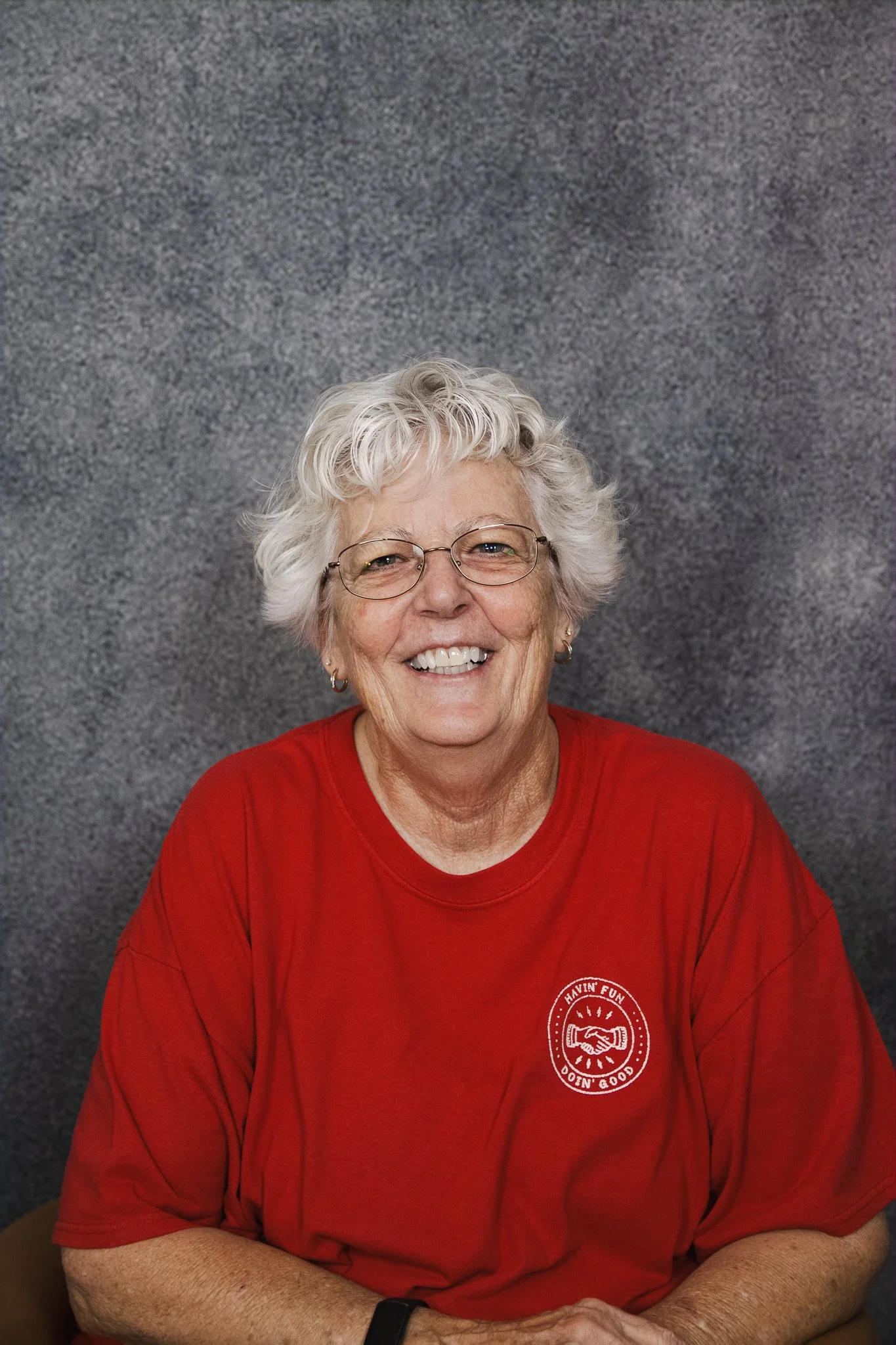 A smiling elderly woman with short curly gray hair and glasses, wearing a red shirt, sitting against a gray background.