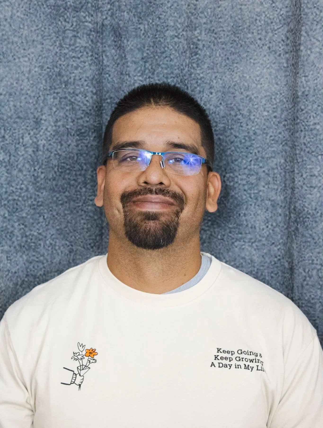 A man with short black hair, wearing glasses, and a goatee, smiling and looking at the camera. He is wearing a white t-shirt with a small illustration of a hand holding a flower, and text that reads "Keep Going & Keep Growing A Day in My Life." The background is a textured blue fabric.