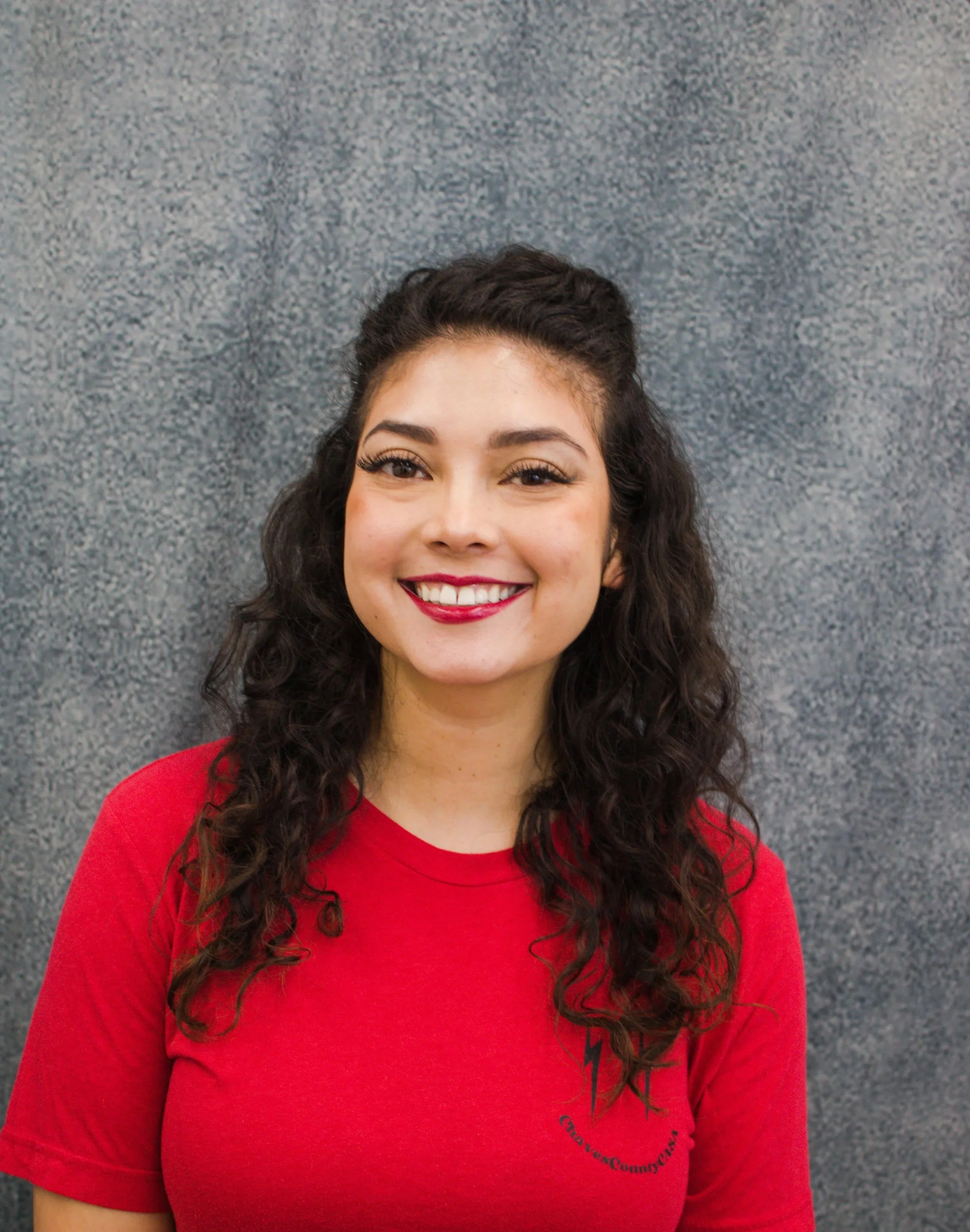A woman with dark curly hair smiling, wearing a red shirt, in front of a gray textured background.