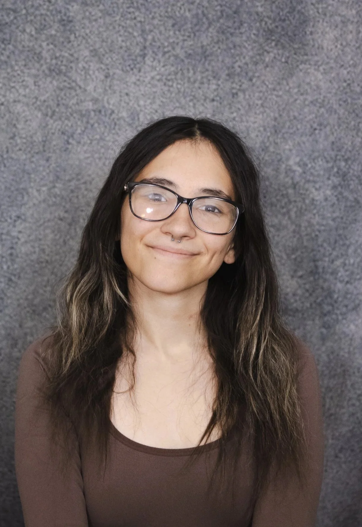 Portrait of a young woman with long dark hair, glasses, a septum piercing, smiling, wearing a brown top, posed against a gray textured background.