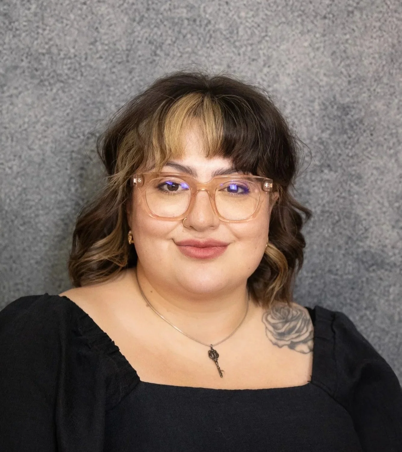 A woman with shoulder-length brown hair with blonde highlights, wearing clear glasses, earrings, a necklace with a key pendant, and a black top. She is smiling and has a tattoo of a rose on her right shoulder. The background is a gray, textured wall.