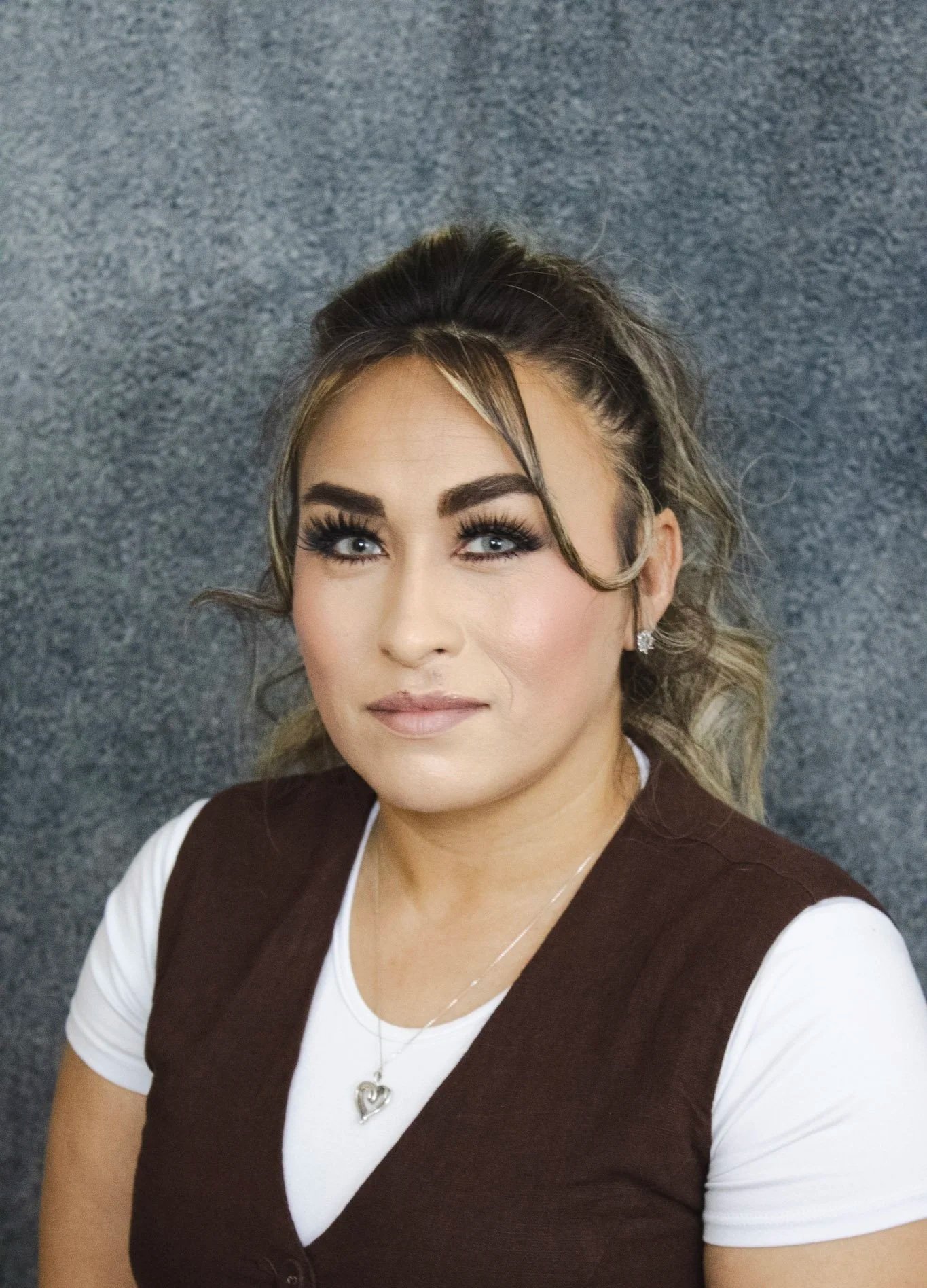 A woman with long eyelashes, styled hair, and diamond earrings, wearing a white shirt and a brown vest, sits against a gray textured background.