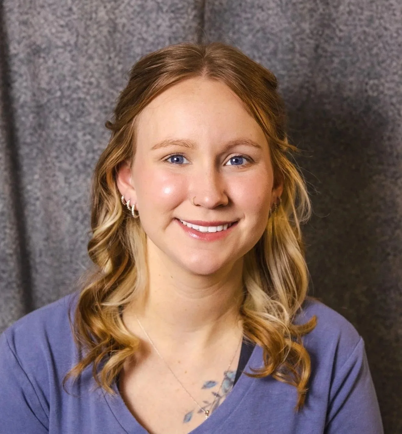 A young woman with wavy blonde hair, blue eyes, and piercings wearing a purple shirt and a delicate necklace, smiling in front of a grey textured background.