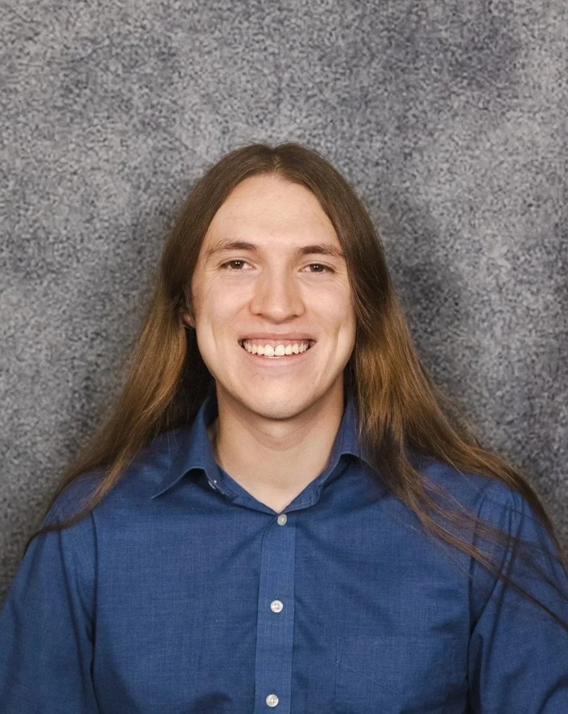 A young man with long brown hair smiling, wearing a blue shirt, in front of a gray textured background.