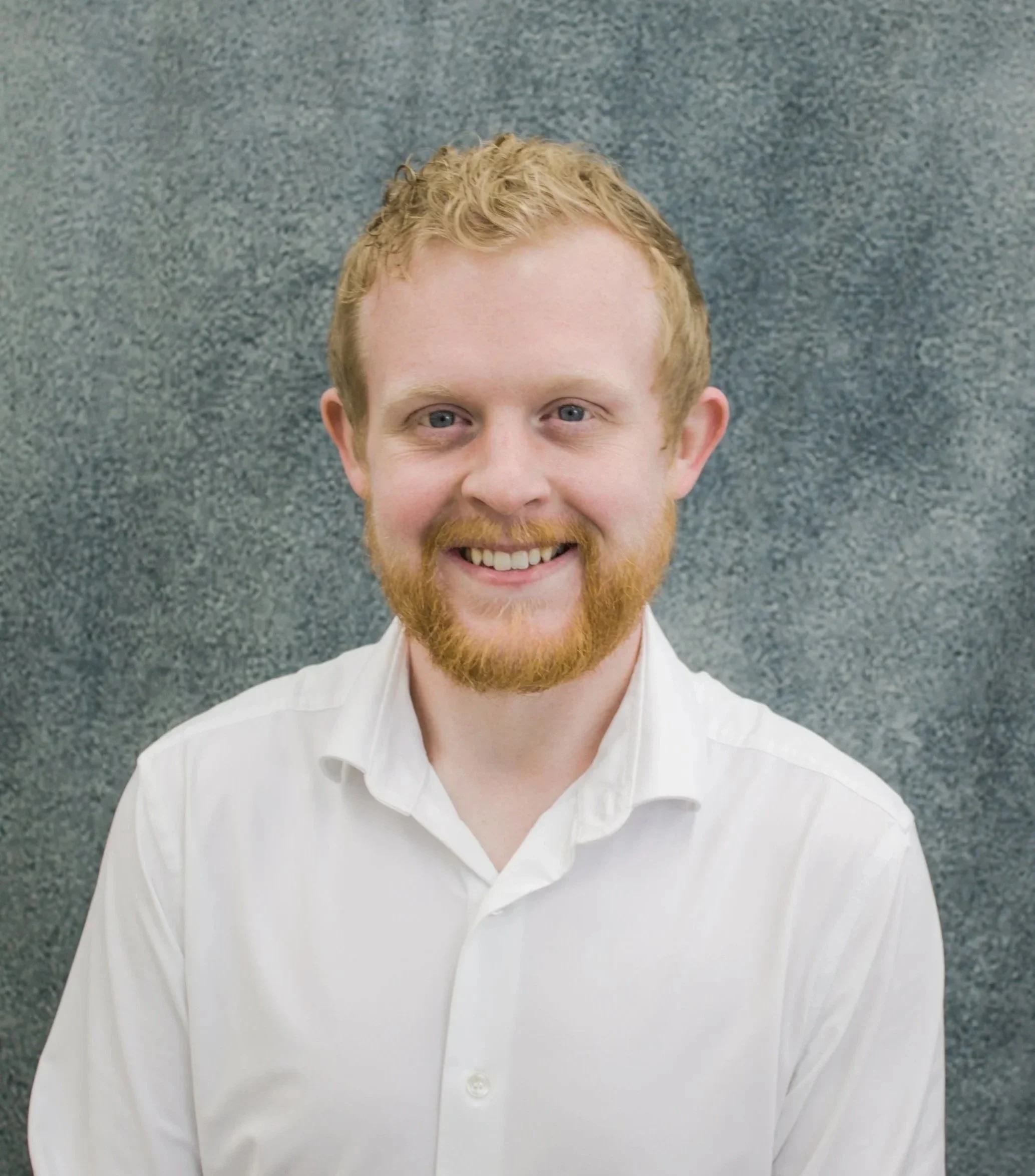 A smiling man with short, curly, ginger hair and a beard, wearing a white shirt, standing in front of a gray textured background.