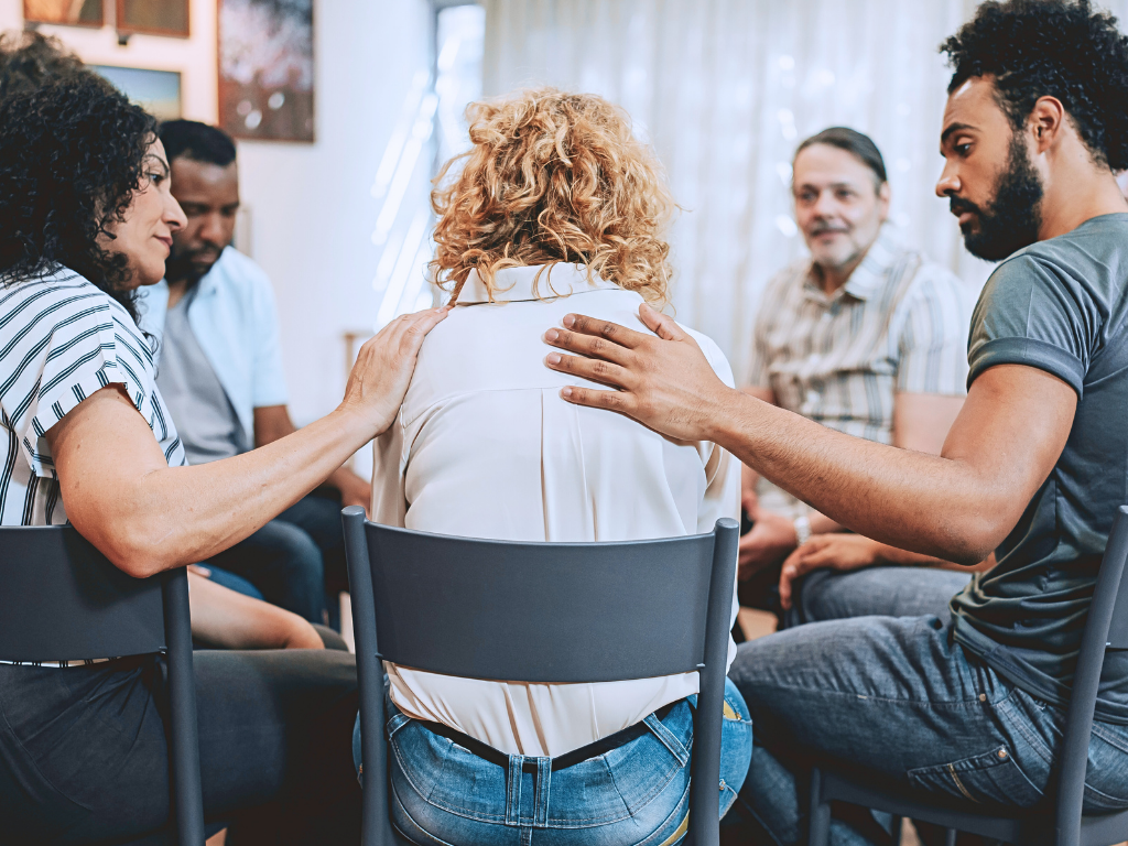Group of people sitting in a circle with one woman with curly blonde hair at the center, receiving comforting support from three others.