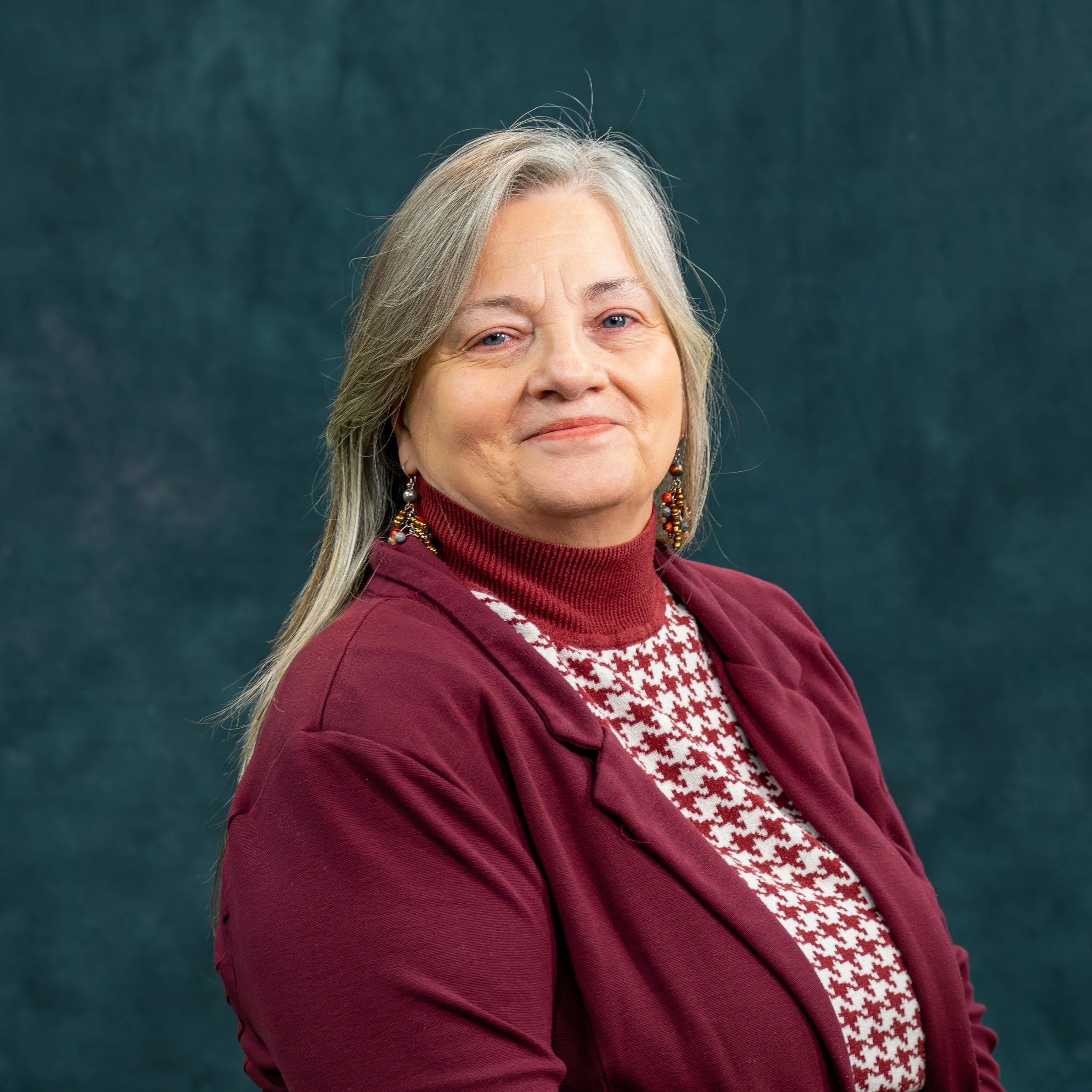 Portrait of an older woman with gray hair, wearing a maroon blazer and patterned sweater, smiling slightly against a dark teal background.