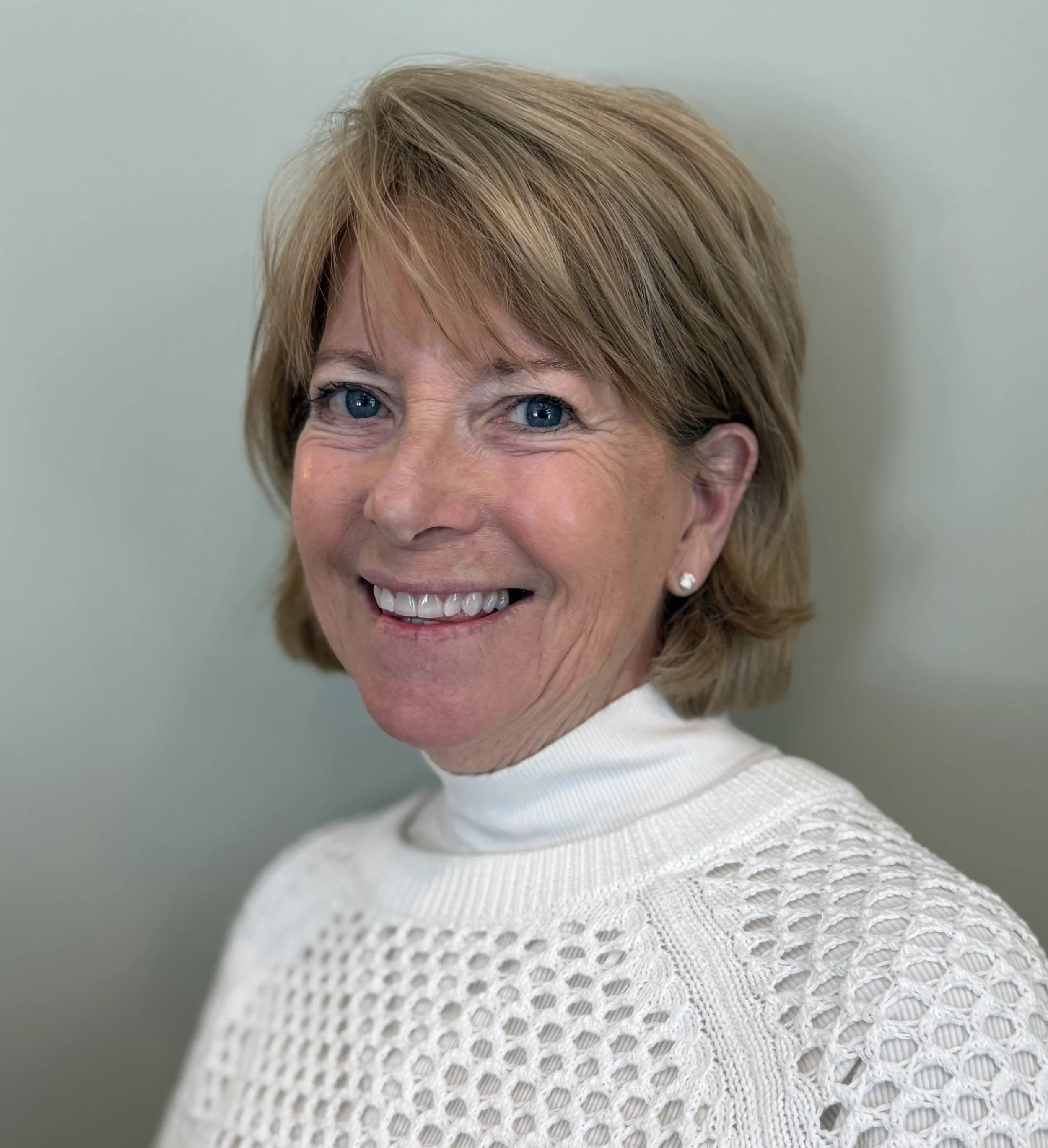 A smiling middle-aged woman with short blonde hair, blue eyes, wearing a white knitted sweater and pearl earrings, standing against a neutral background.