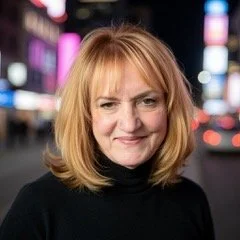 A woman with shoulder-length reddish blonde hair, smiling, standing outdoors at night in an urban setting with blurred city lights in the background.
