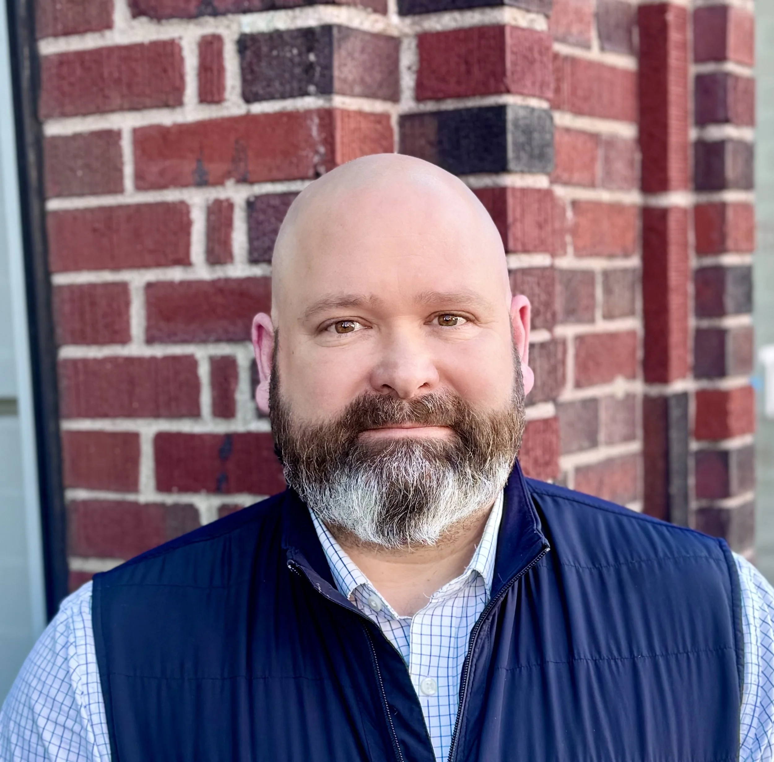 Headshot of a bald man with a beard, wearing a white checkered shirt and a navy vest, standing outside in front of a red brick wall.