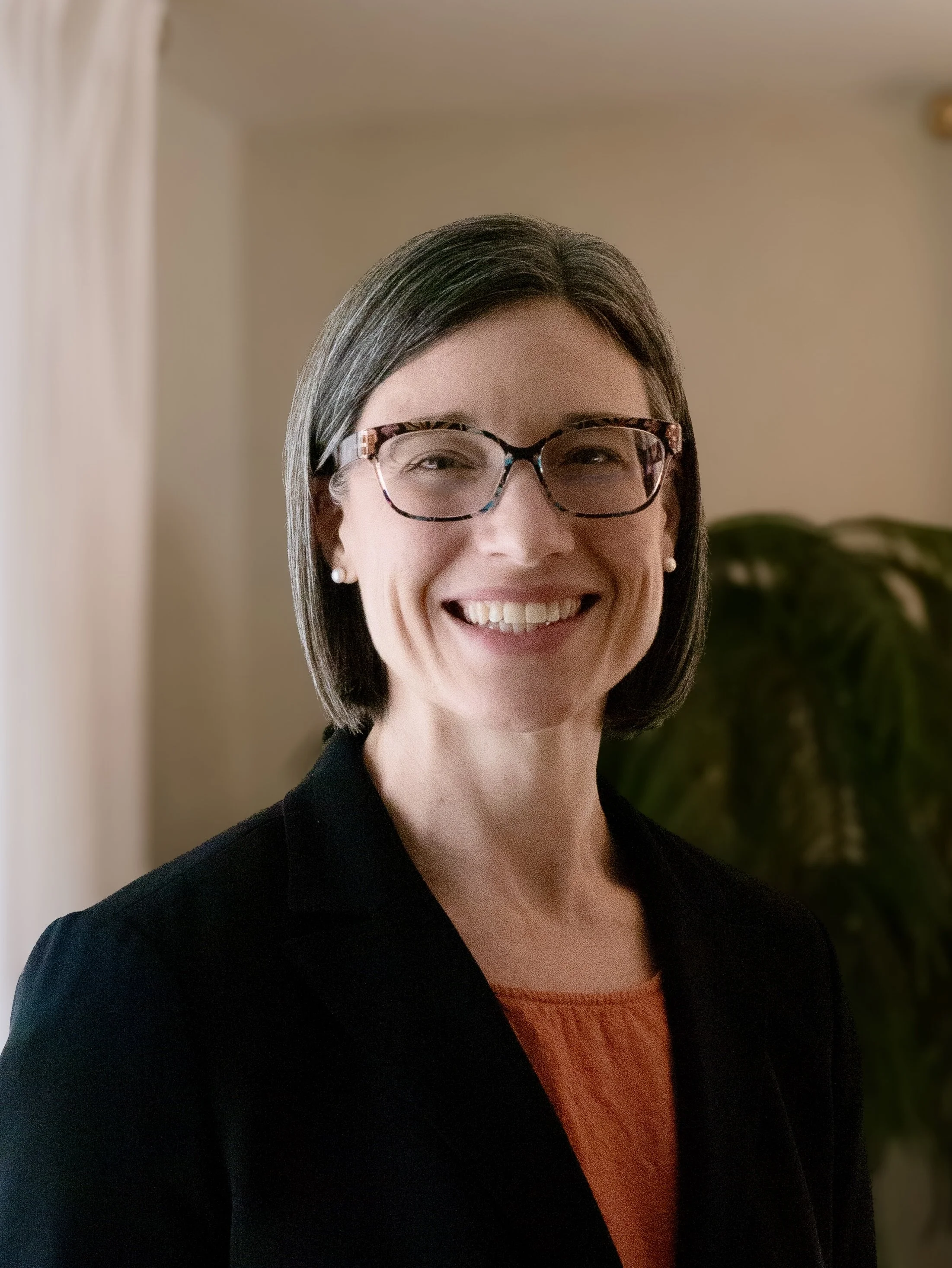 A woman with short dark hair, wearing glasses with tortoiseshell frames, pearl earrings, a black blazer, and an orange top, smiling indoors with a plant in the background.