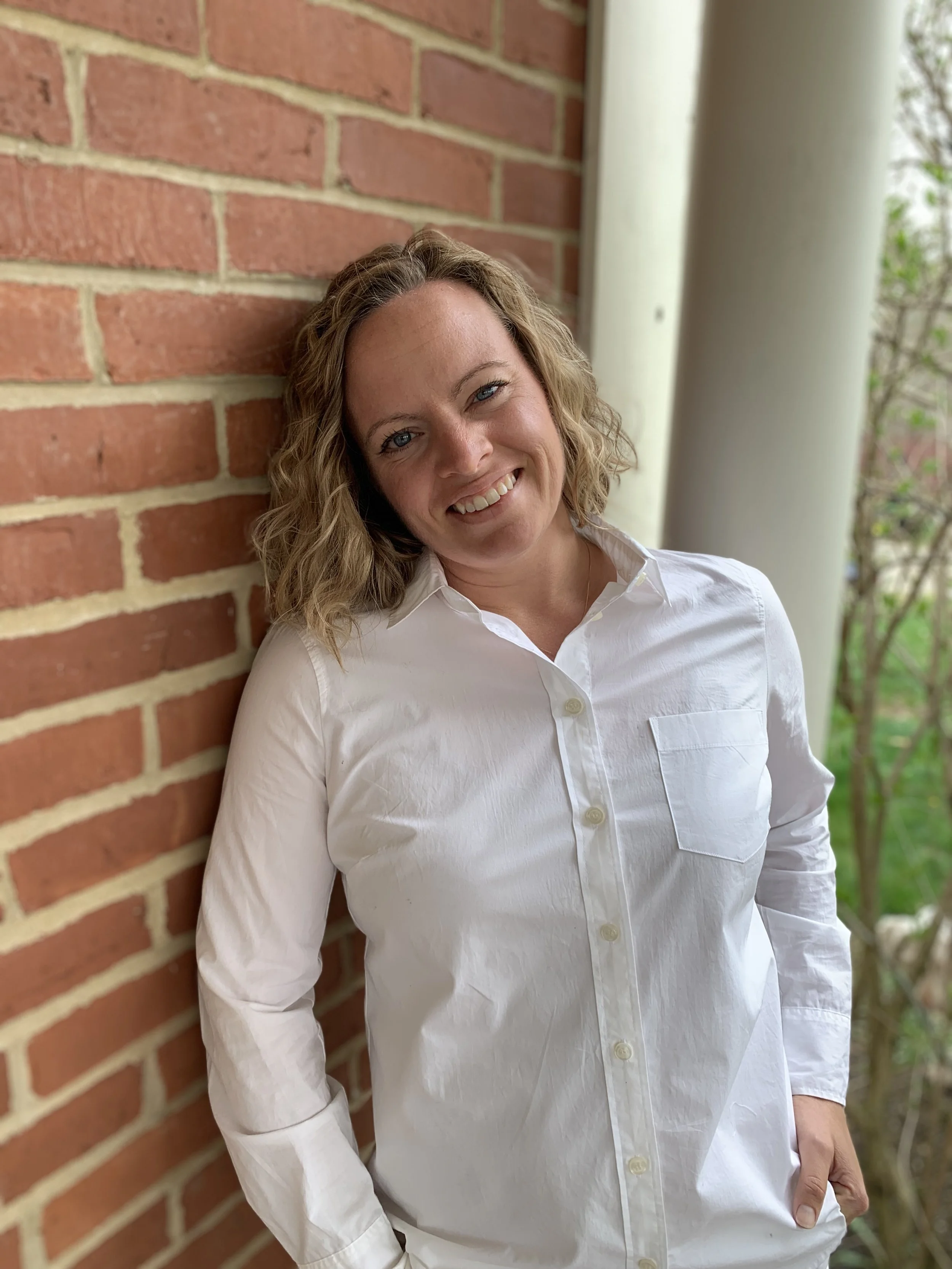 A woman with curly blonde hair in a white button-up shirt leaning against a brick wall outdoors, smiling at the camera.