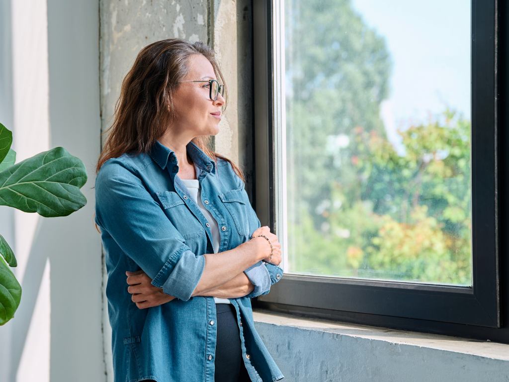 A woman with glasses wearing a blue denim shirt looking out of a window with trees outside.