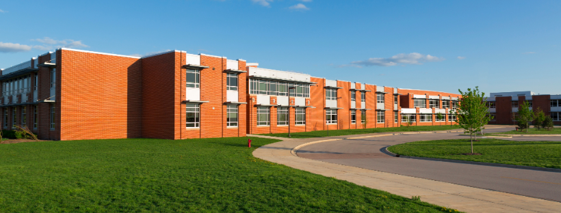 A modern, brick educational building with multiple windows, located next to a curved road and green grassy area with trees under a clear blue sky.