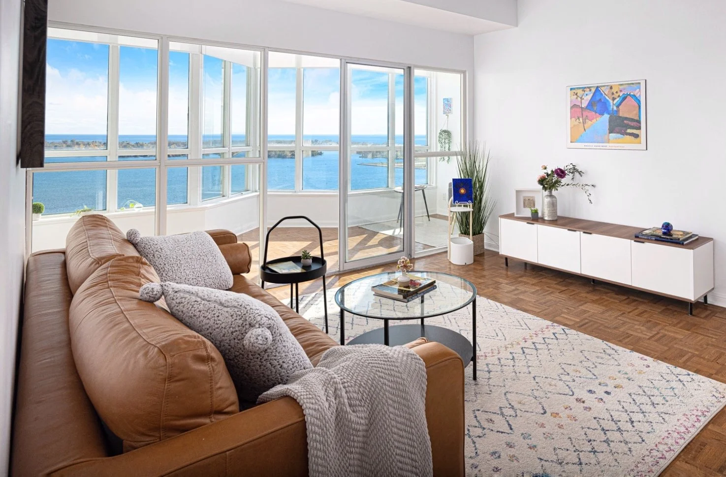 Bright living room with large windows overlooking water, featuring a tan leather sofa with textured pillows, a round glass coffee table, white and wood sideboard with decorative items, and a balcony with outdoor seating, all illuminated by natural light.