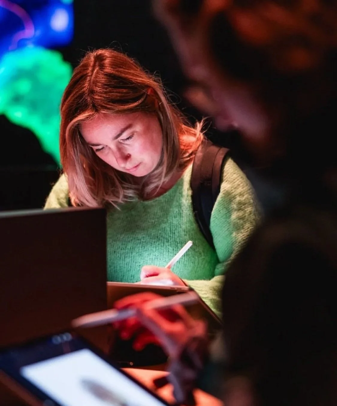 A woman with shoulder-length brown hair writing with a pen at a table in a dimly lit setting with neon lighting in the background.