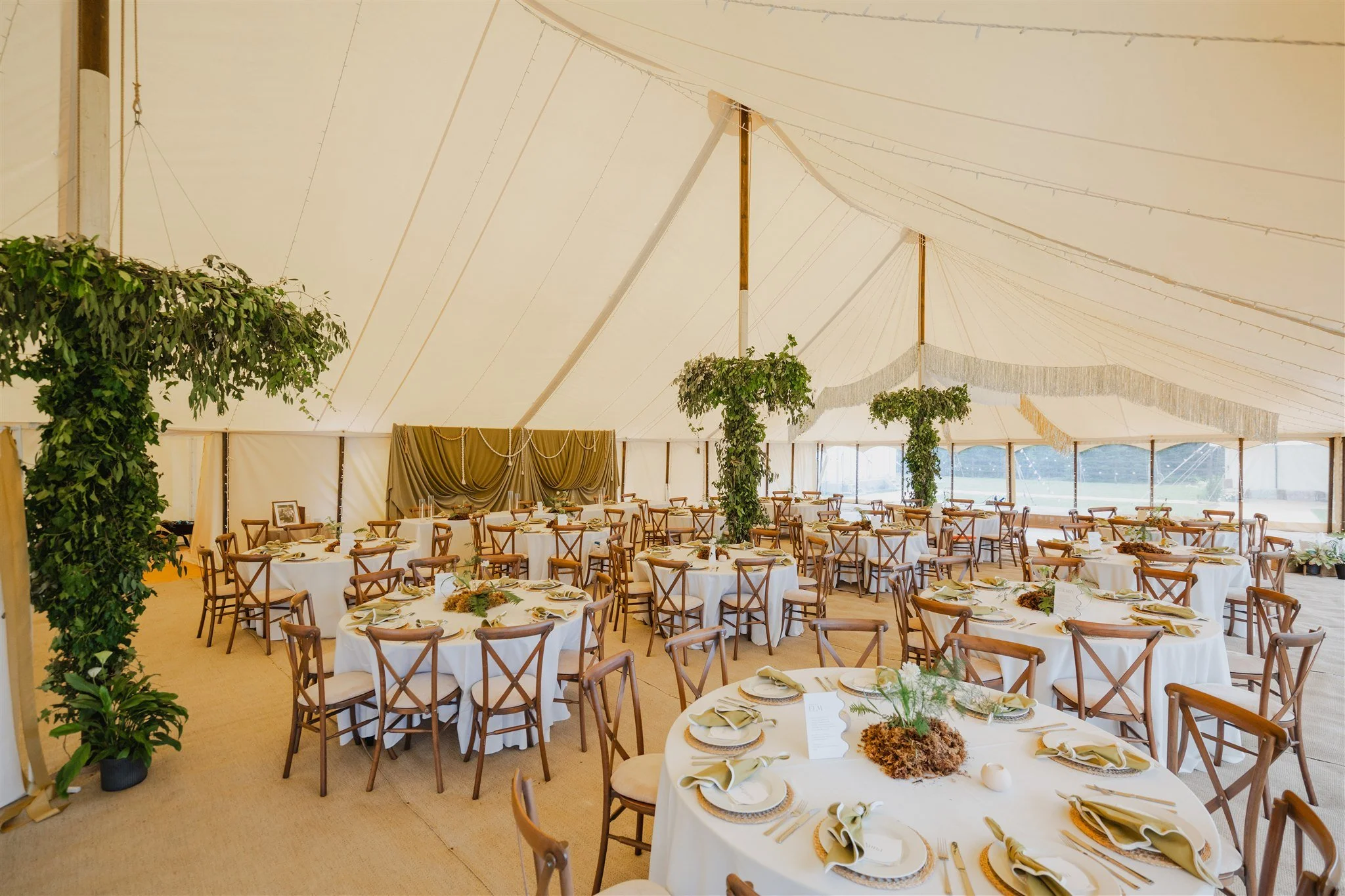 Interior of a decorated event tent with round tables covered in white tablecloths, set for a meal with plates, napkins, and silverware, and accented with floral centerpieces and greenery.