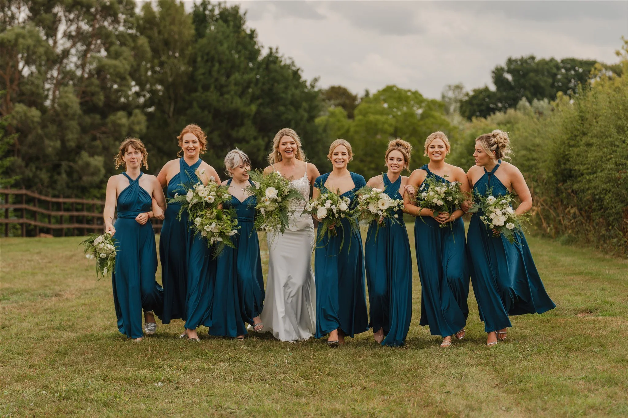 Group of nine women walking outdoors in a park, dressed in blue bridesmaid dresses with one woman in a white wedding gown, holding bouquets of white flowers, with trees and cloudy sky in the background.