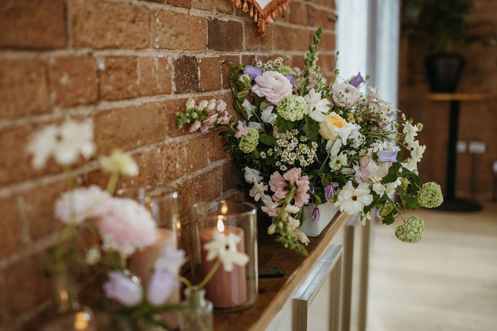 A floral arrangement with white, pink, and purple flowers on a wooden surface against a brick wall, with candles and blurred flowers in the foreground.