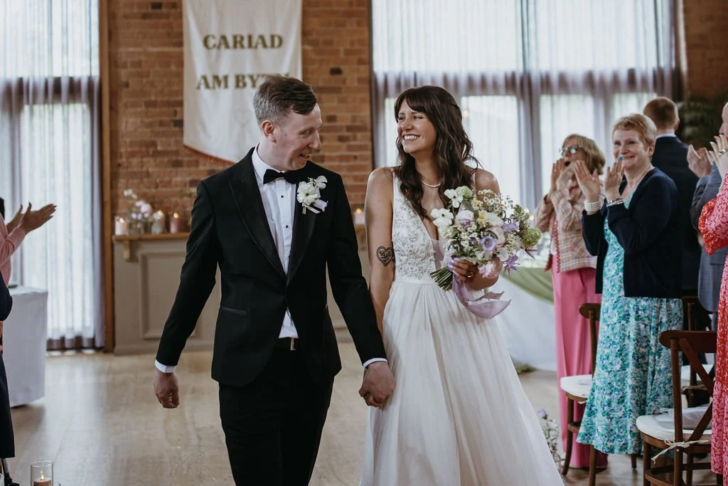 Bride and groom holding hands and smiling at each other in a wedding ceremony, with guests clapping in the background.