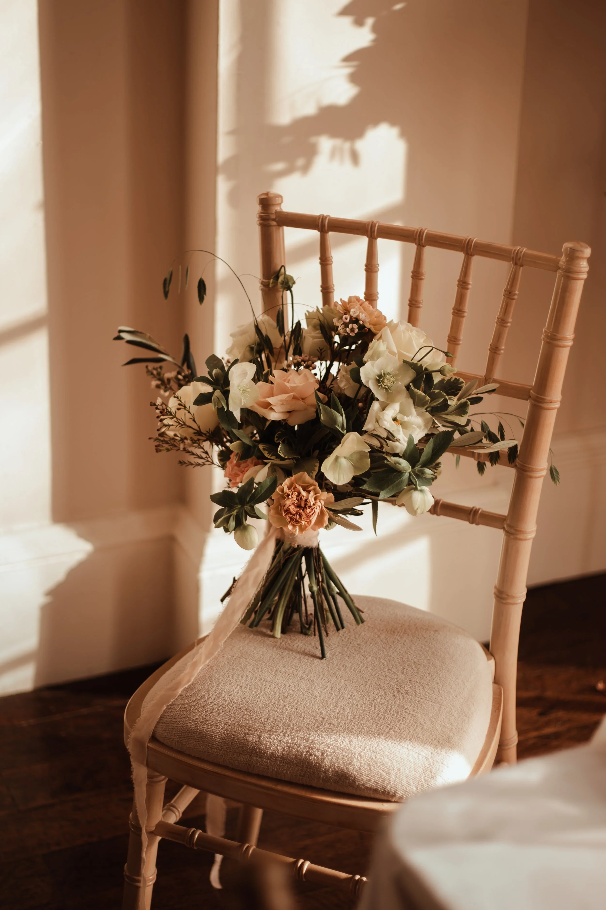 A bouquet of white and blush pink flowers resting on a beige cushion chair, with warm sunlight casting shadows on the wall behind.
