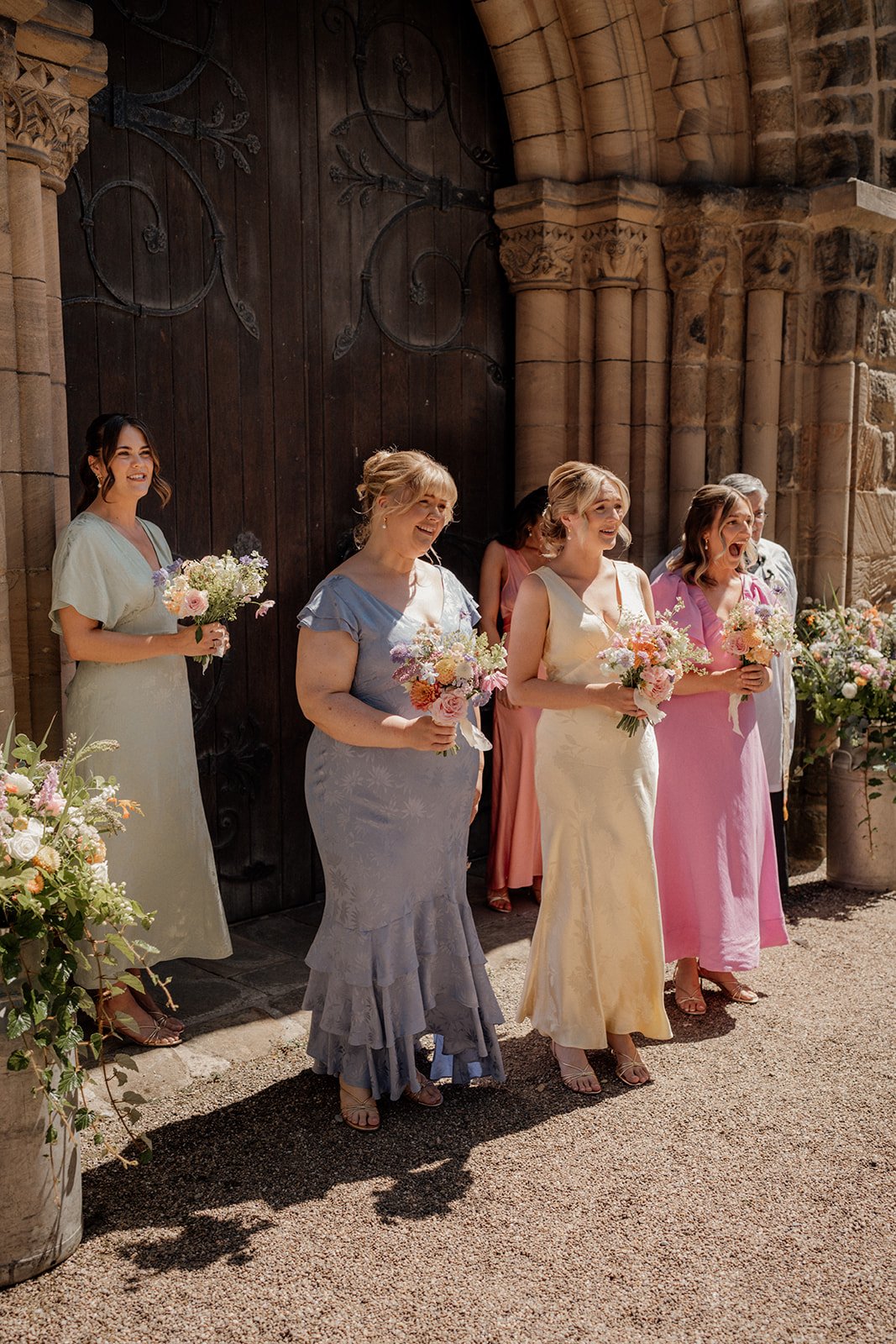 Women standing outside a stone church, dressed in colorful dresses, holding bouquets of flowers, smiling during a wedding ceremony.