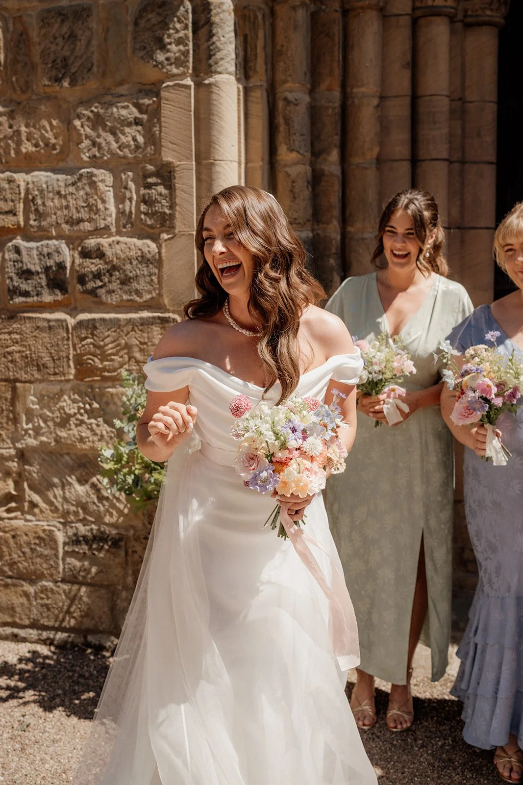 Bride with long brown hair in a white gown holding a bouquet of flowers, smiling and laughing outside with bridesmaids in pastel dresses behind her.