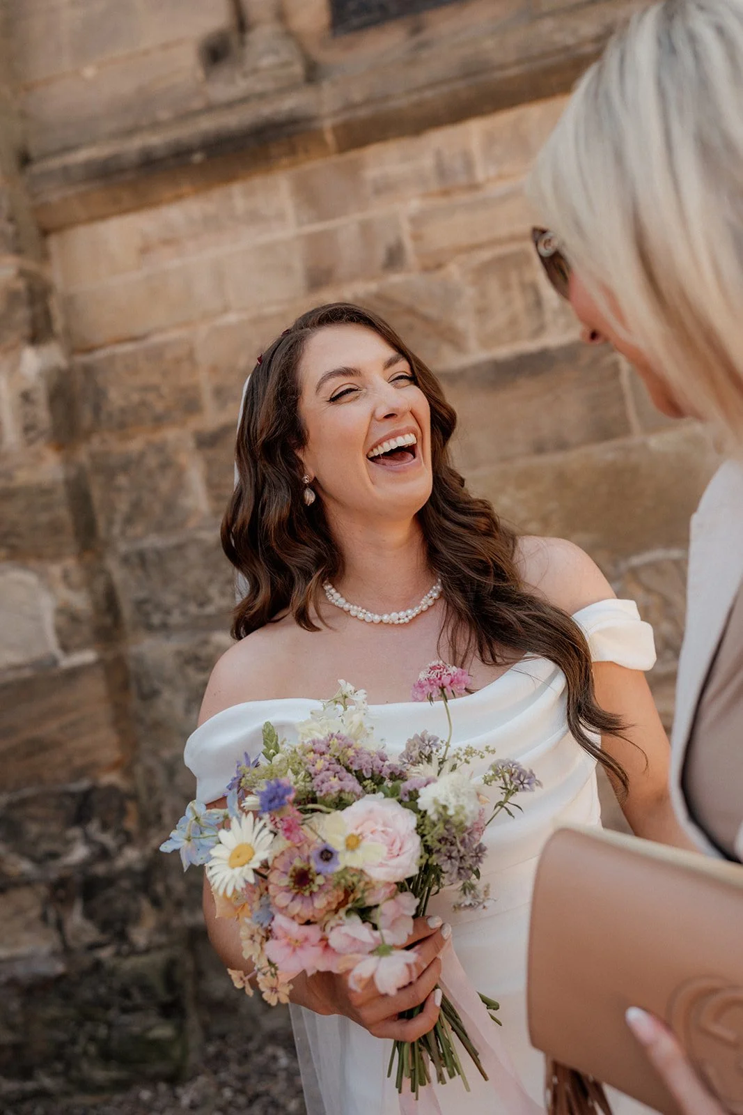 A bride with long dark hair, wearing a pearl necklace and an off-shoulder white wedding dress, holding a bouquet of pink, white, and purple flowers, is laughing joyfully while looking at her partner against a stone wall background.